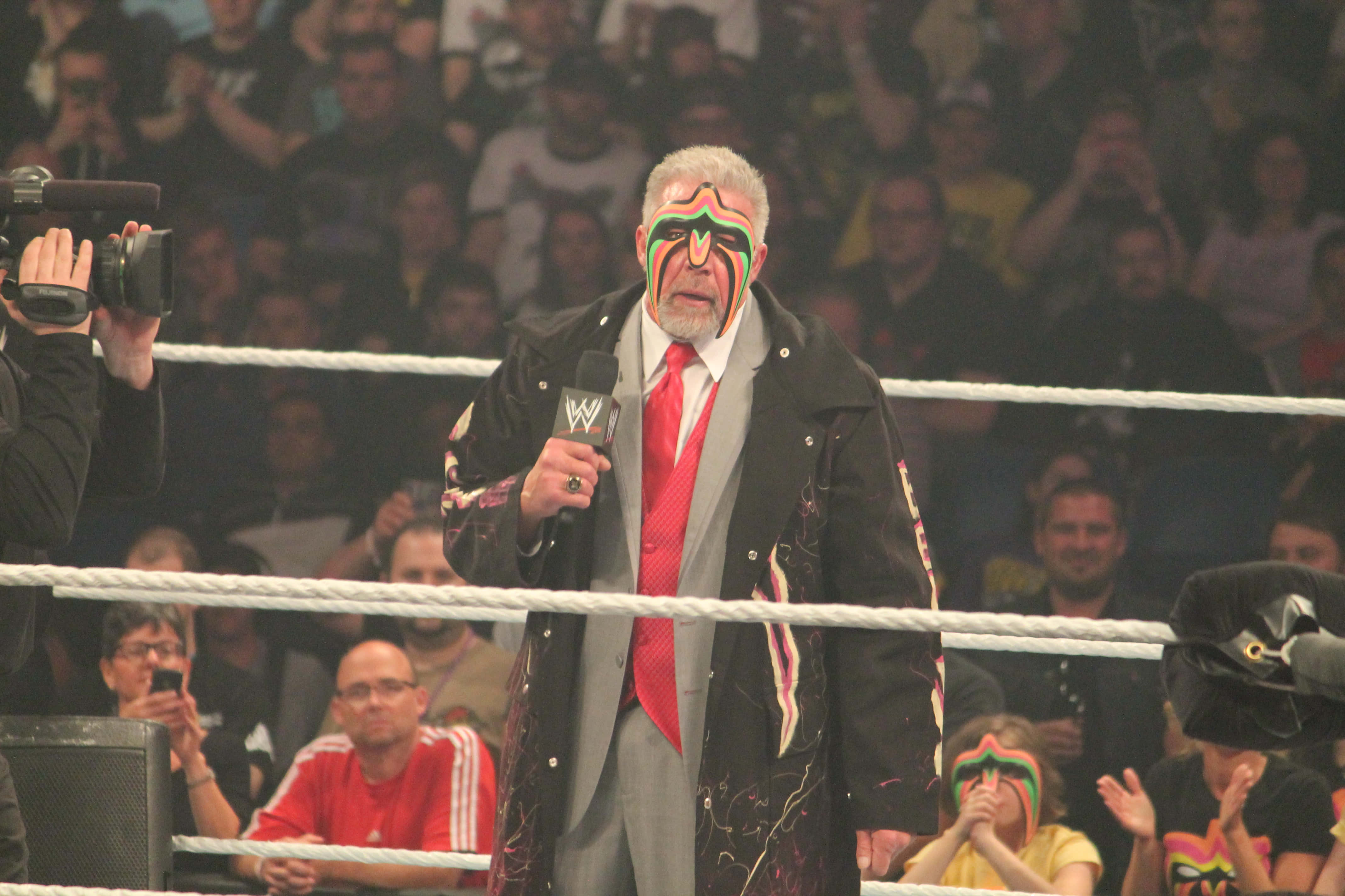 Ultimate Warrior 在 WWE Raw 2014 現場演出 壁紙 — 摔角手臉上塗著彩色顏料，站在擂台上拿著麥克風，觀眾熱切觀看。