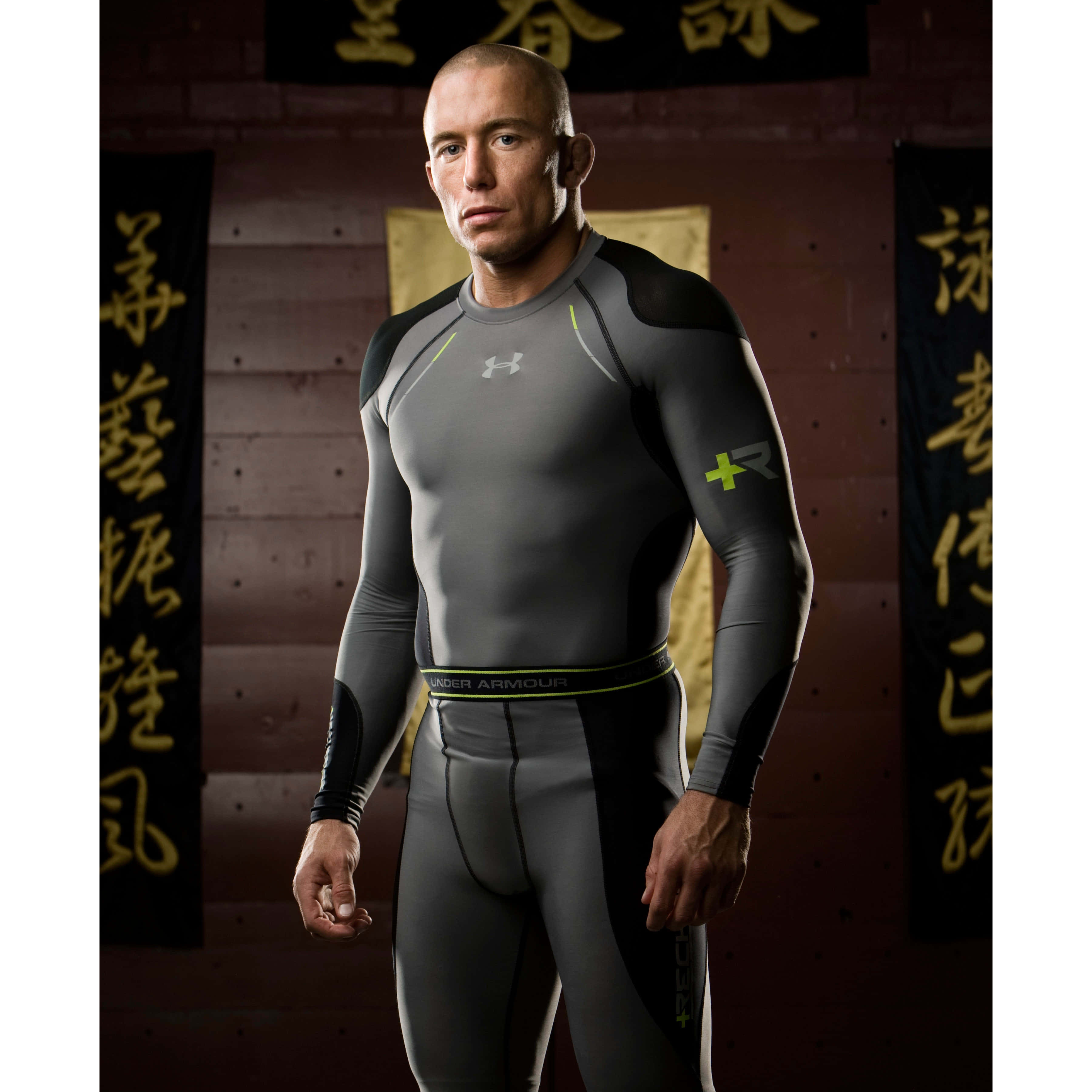 穿上 Under Armour，展現與感受運動員風範 壁紙 — 一個肌肉發達的男人穿著灰色和黑色的Under Armour壓縮裝備，自信地站在一面裝飾著中國書法橫幅的磚牆前。