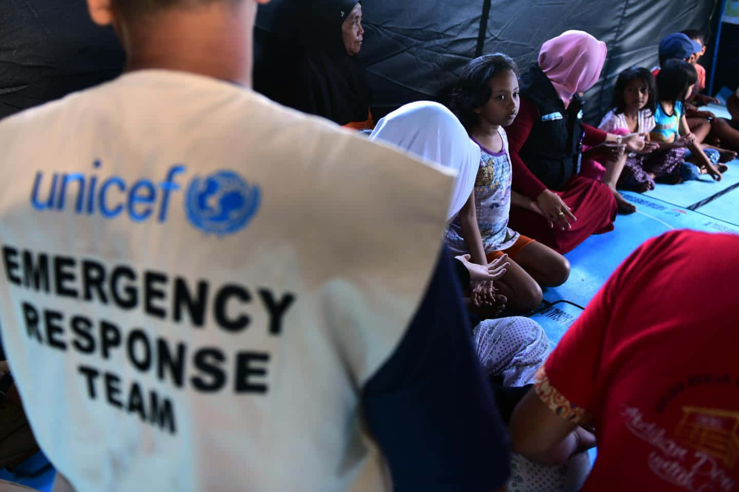 UNICEF人道救援行動 壁紙 — 聯合國兒童基金會緊急應變人員在深色布料牆壁下的臨時避難所中照顧坐在墊子上的兒童。