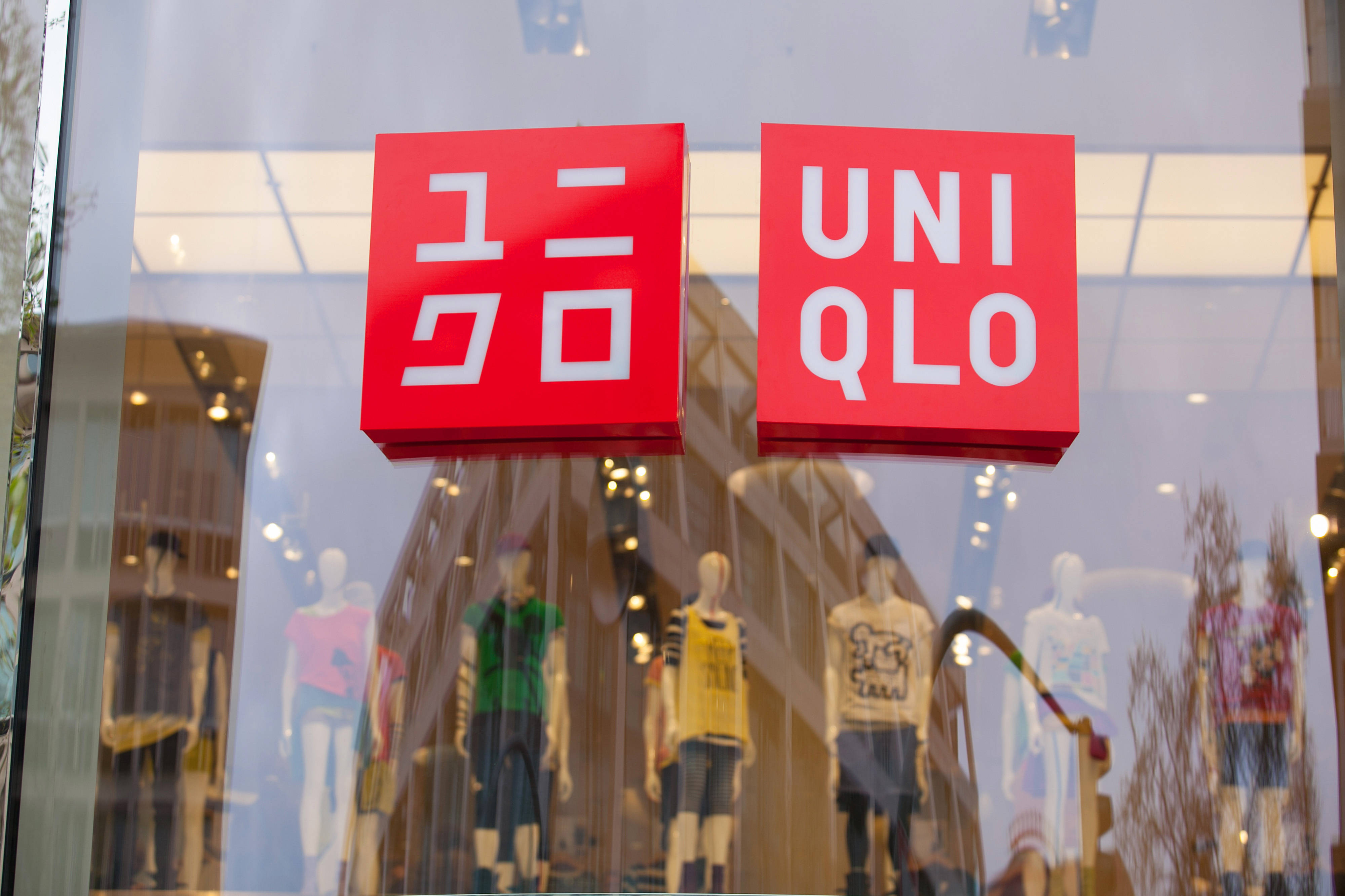 UNIQLO 紅色標誌 壁紙 — 一個鮮紅色的Uniqlo標誌懸掛在玻璃櫥窗內穿著色彩繽紛休閒服飾的人體模型上方。
