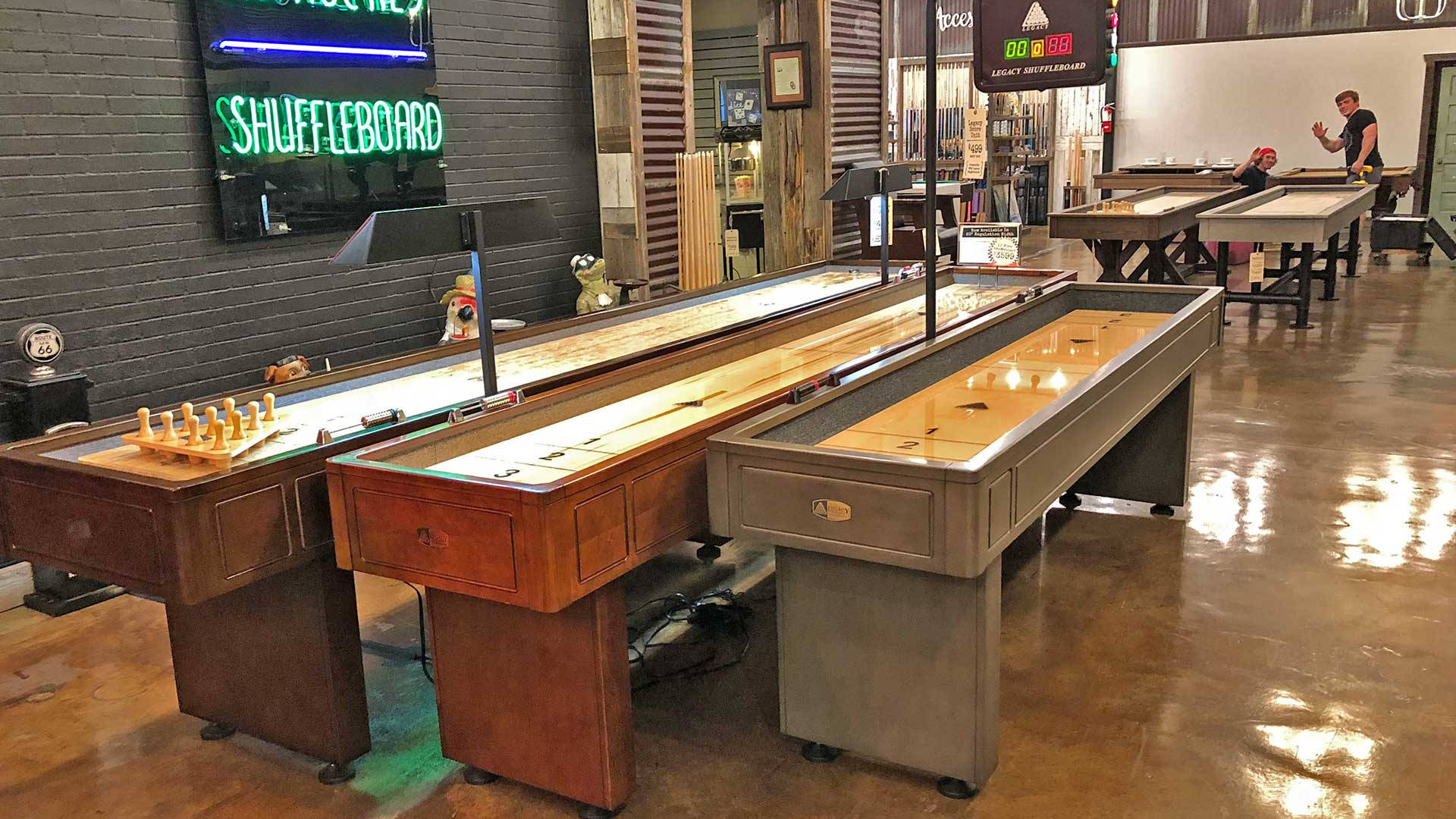 Universal Billiards 木質 Shuffleboard 桌 壁紙 — 三張帶有木製軌道和圓盤的沙狐球桌，放置在帶有霓虹燈招牌和拋光混凝土地板的復古遊戲廳中。