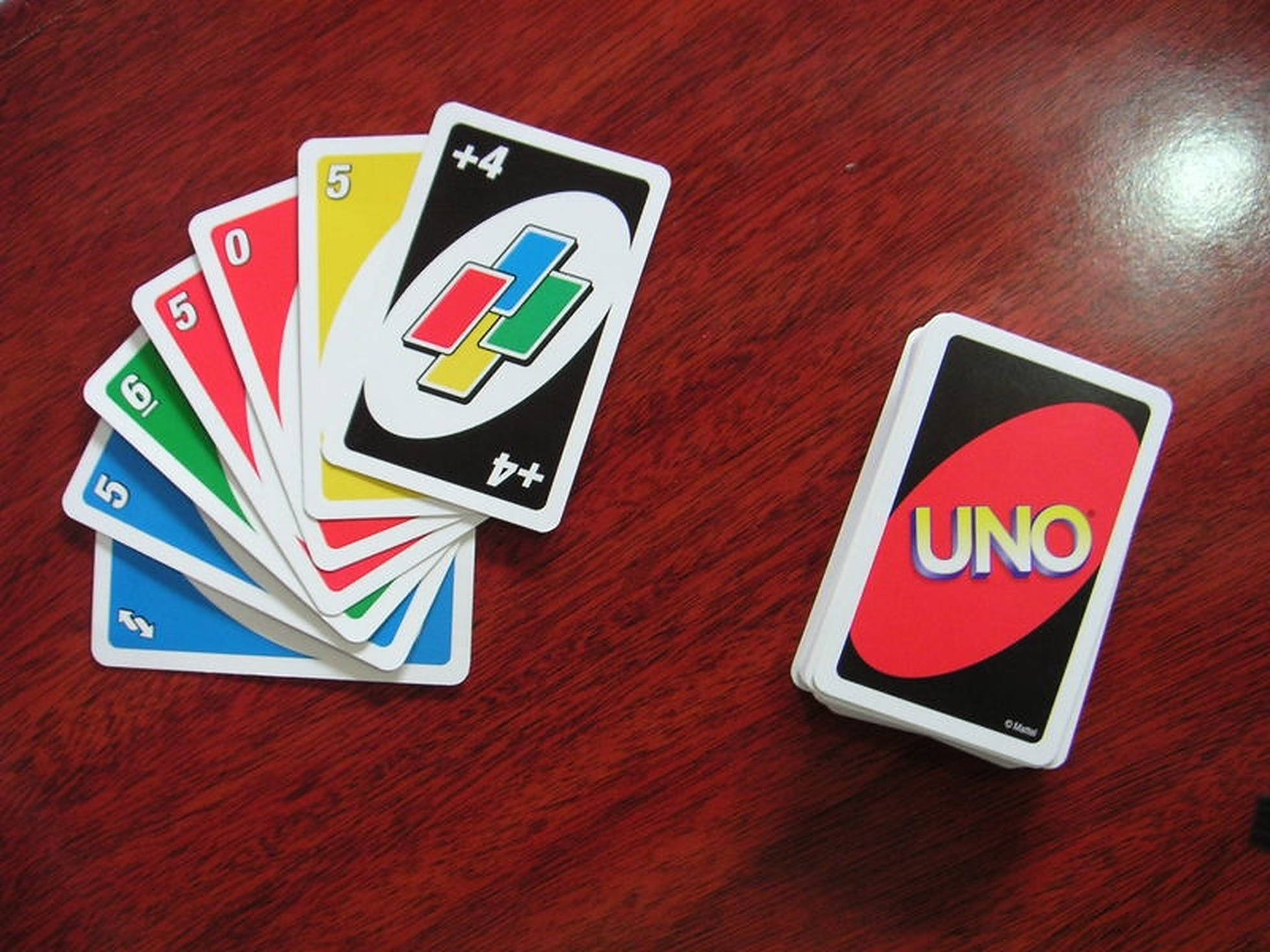Uno 卡牌組 壁紙 — 色彩繽紛的UNO卡牌扇形散開，放在一張拋光紅木桌上，旁邊有一副牌。
