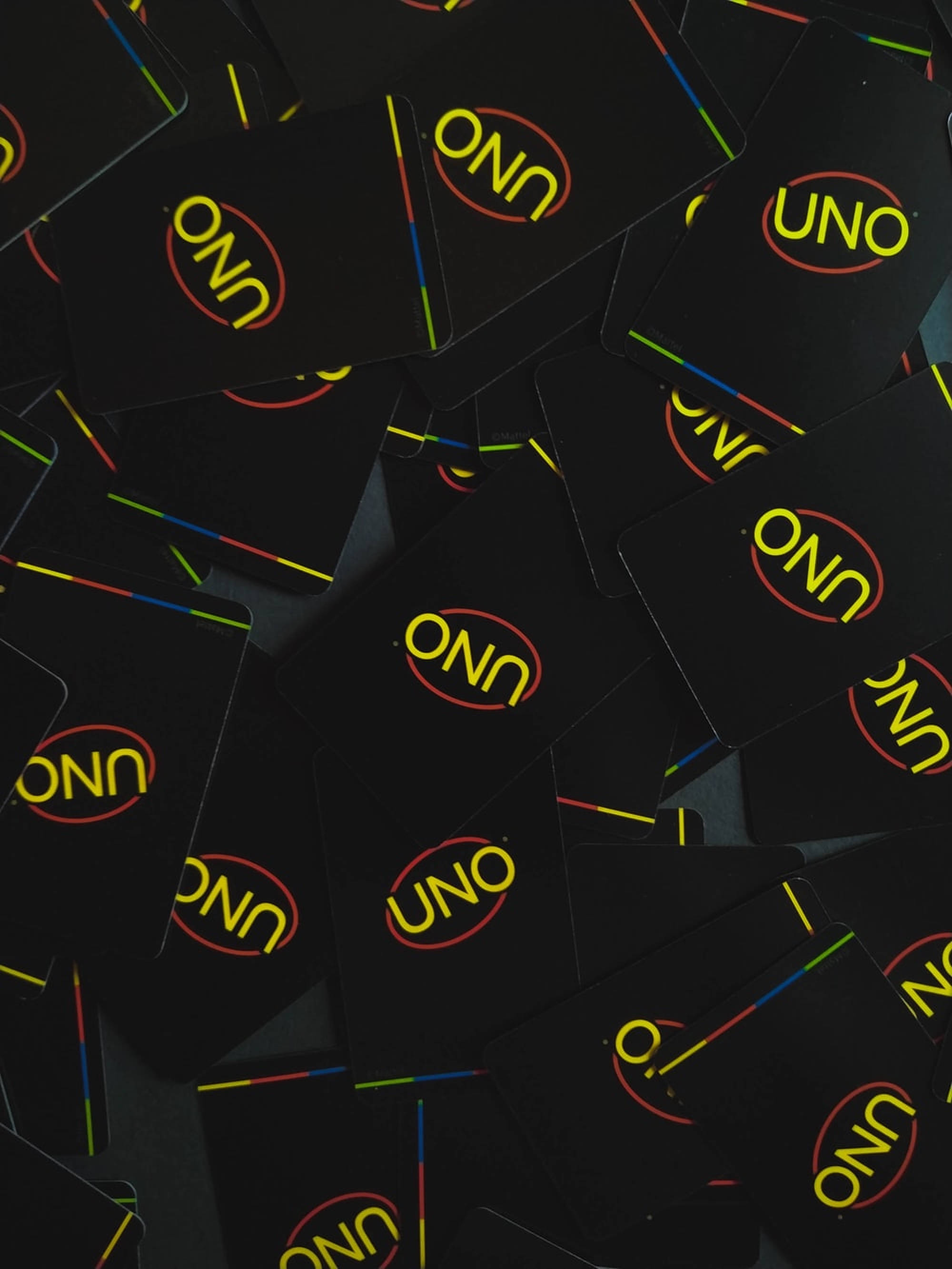 Uno 卡牌組極簡設計 壁紙 — 黑色UNO牌散落在深色表面上的特寫，帶有黃色標誌和彩色條紋。