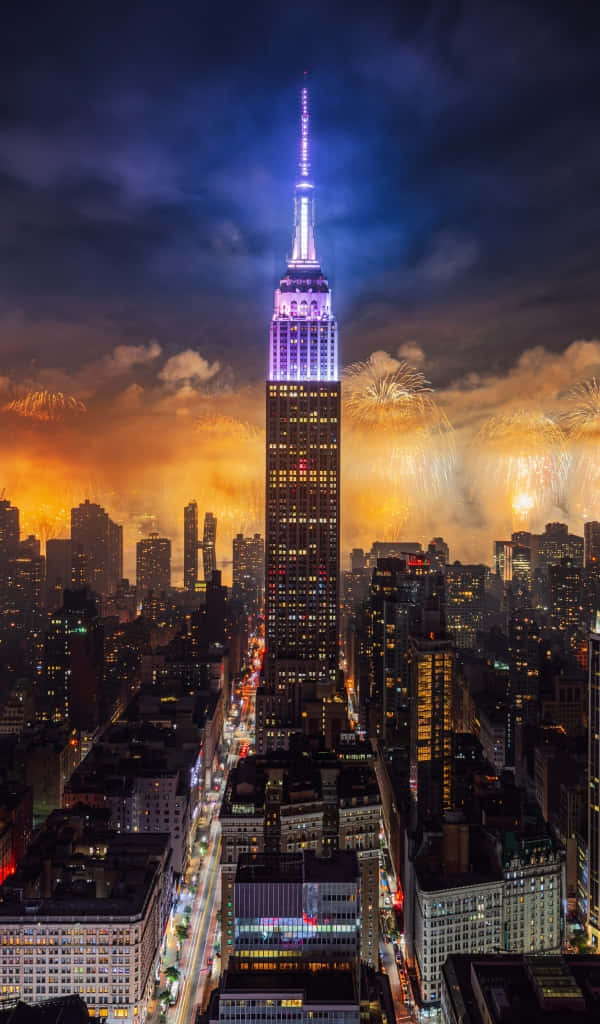 Unsplash 精選紐約市璀璨天際線 壁紙 — 帝國大廈在黑暗的天空中發出紫色和藍色的光芒，煙火在紐約市上空以金色爆發綻放。