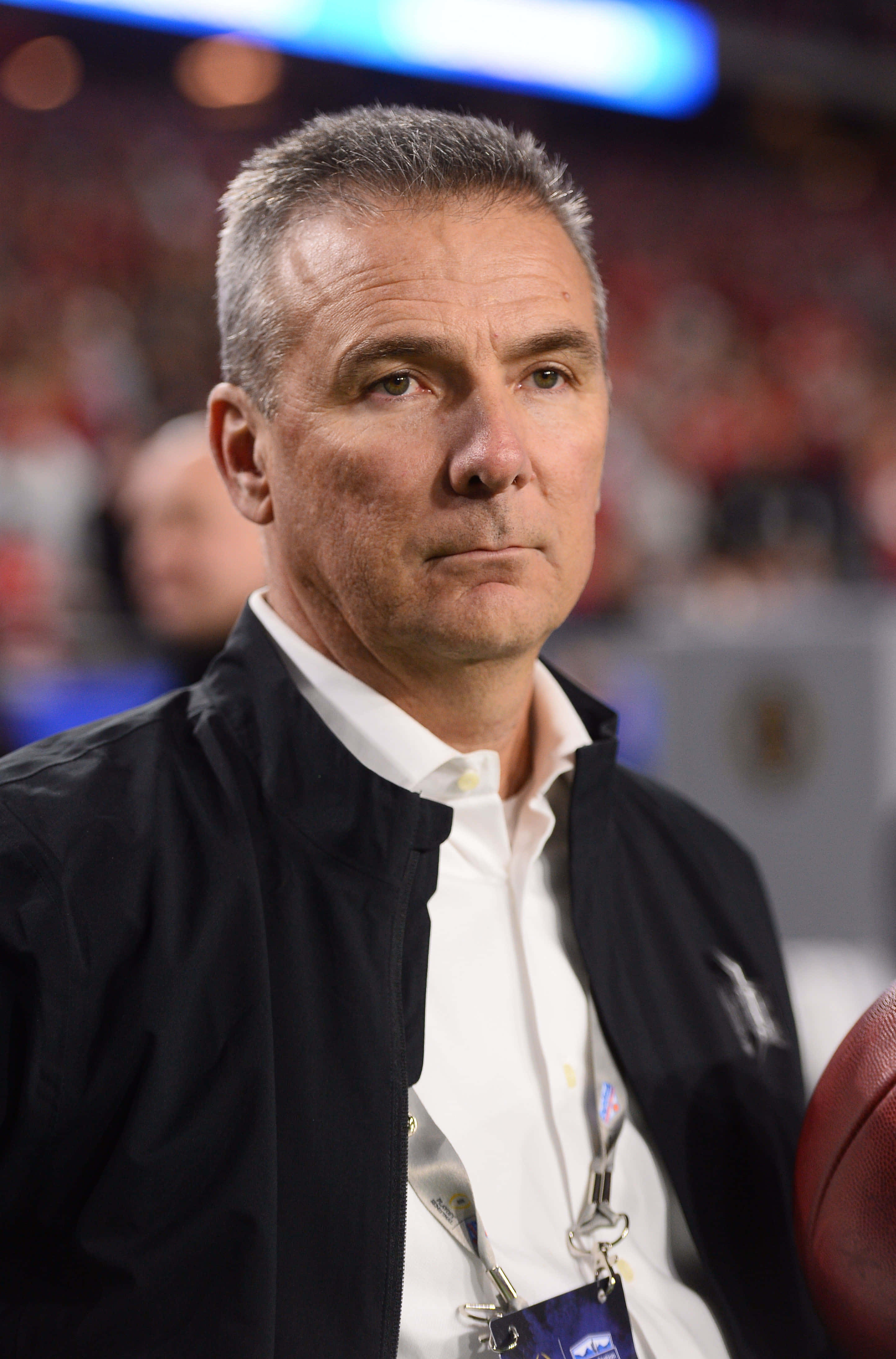 Urban Meyer — 勝利的足球教練 壁紙 — 一個專注的中年男子穿著黑色外套，在燈光明亮的體育館裡拿著一個足球，身後是模糊的紅色座位。
