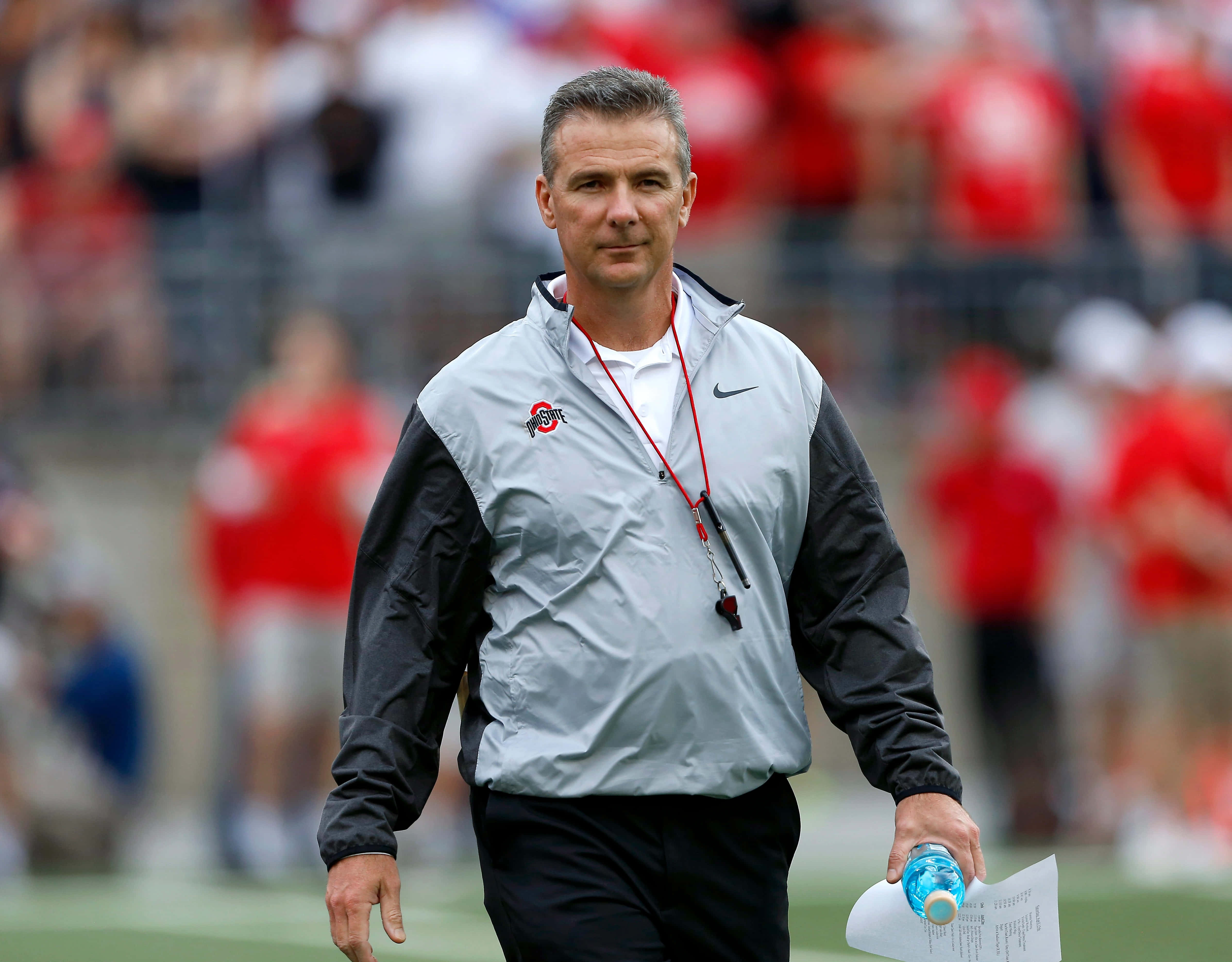 Urban Meyer 帶來對足球的熱情與卓越 壁紙 — 一位專注的足球教練穿著灰色和黑色外套，拿著水樽和文件在綠色球場上行走。