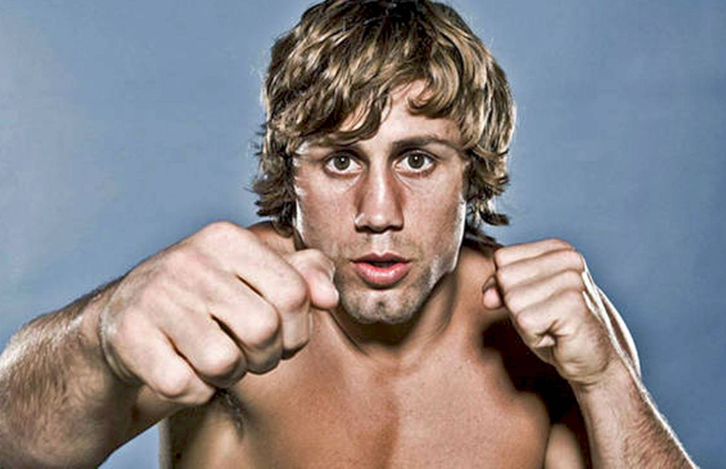 Urijah Faber 擊拳姿勢 壁紙 — 一名赤裸上身的男性拳擊手，頭髮凌亂，在純藍色背景下，握緊拳頭擺出戰鬥姿勢，目光銳利地直視前方。