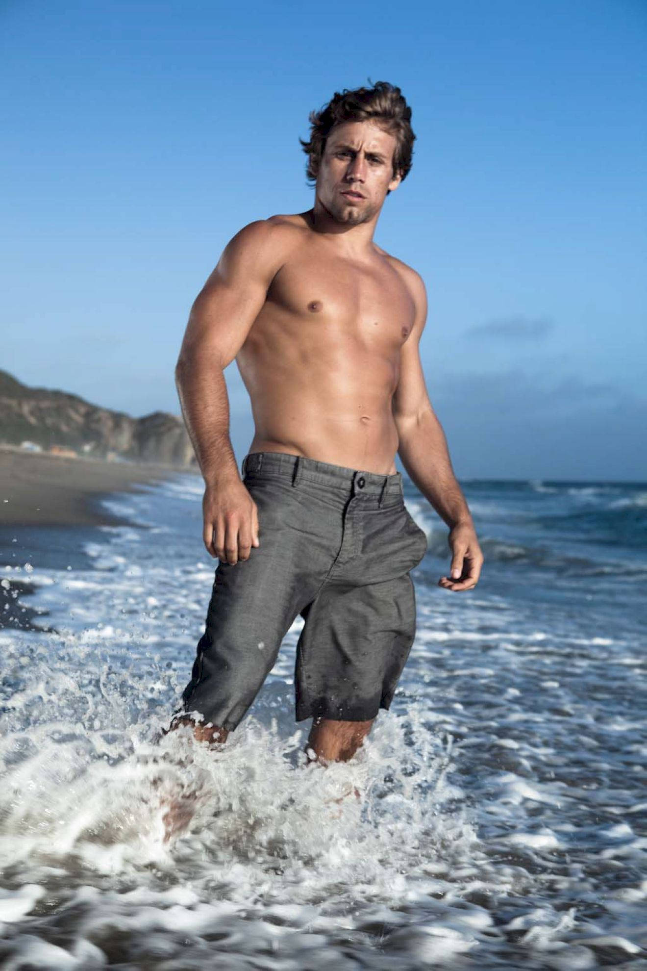 Urijah Faber 在海灘 壁紙 — 一個肌肉發達的男人在陽光明媚的海灘上穿過海浪，背景是懸崖。