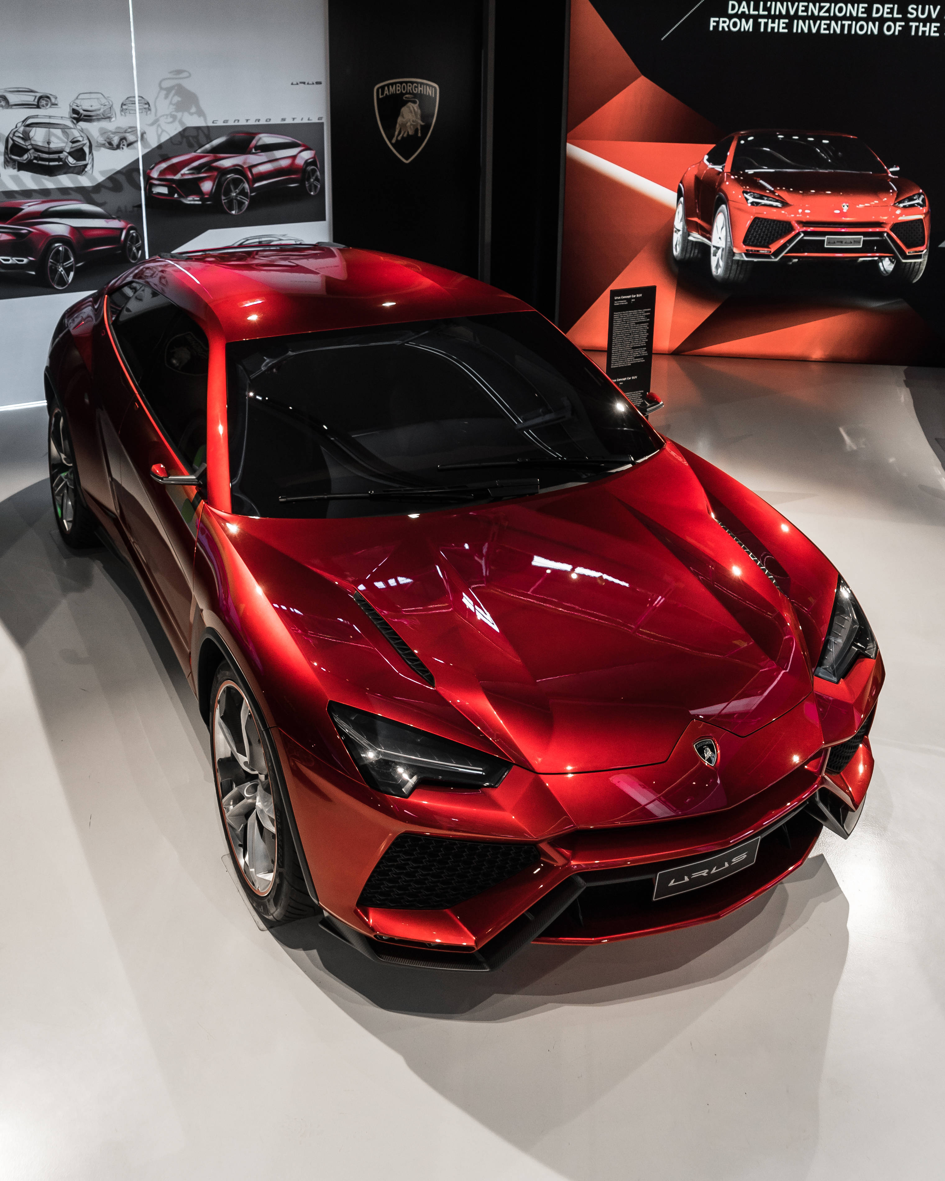 紅色蘭博基尼Urus豪華SUV 壁紙 — 一輛光澤的紅色林寶堅尼 Urus SUV 在明亮燈光下，於一個時尚現代、帶有品牌牆壁和概念藝術的陳列室中閃閃發光。