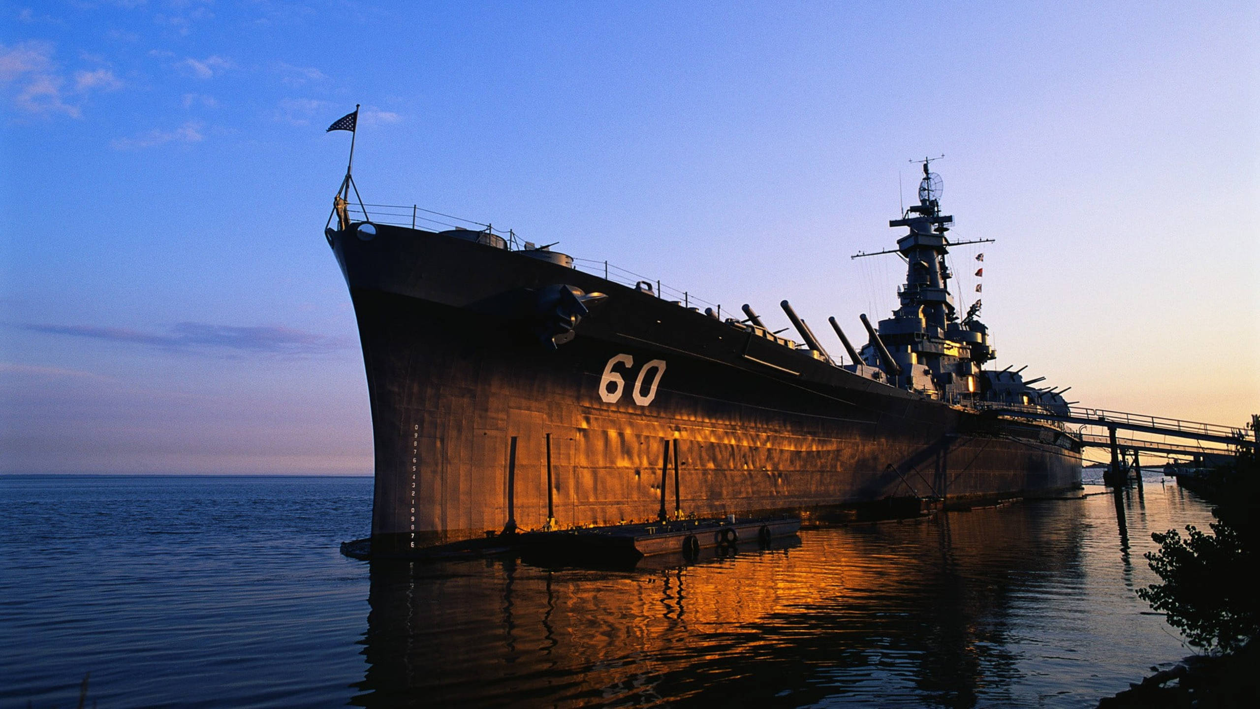 USS 阿拉巴馬號戰艦日落 壁紙 — 一艘巨大的戰艦，編號60，被金色的夕陽照亮，靜止在平靜的水面上，倒映出它的輪廓，映襯著暮色天空。