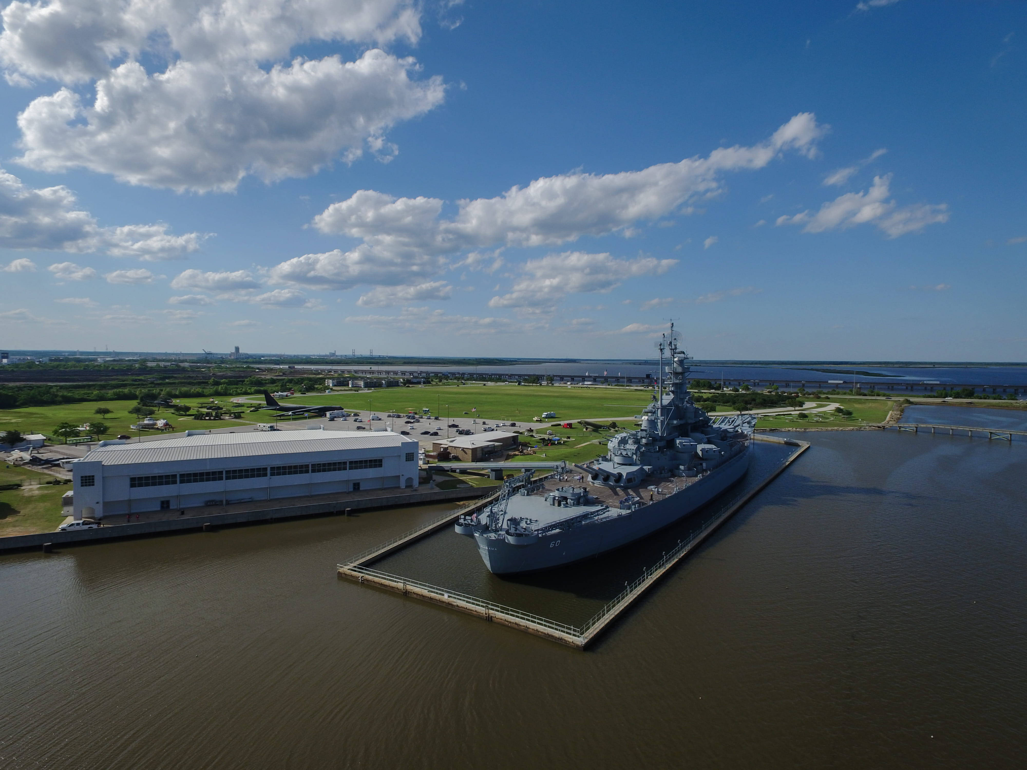 USS 阿拉巴馬號戰艦空中俯瞰 壁紙 — 一艘大型灰色戰艦停泊在碼頭的航拍圖，背景是明亮的藍天和白雲點綴。