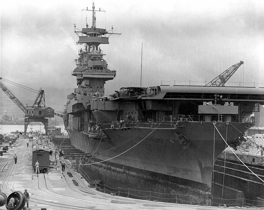 USS約克鎮於珍珠港準備出擊 壁紙 — 一艘巨大的航空母艦停泊在造船廠，船體周圍有起重機和工人，天空多雲。