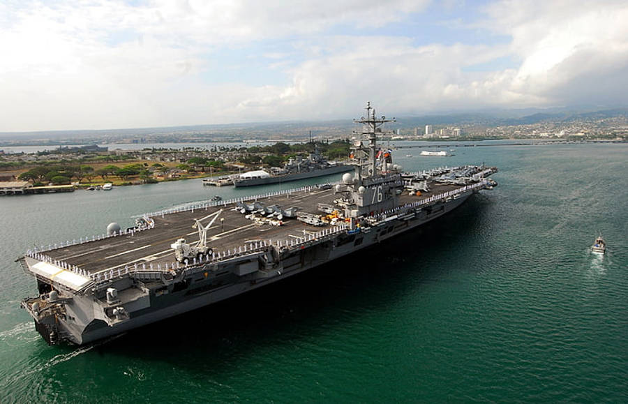 USS羅納德·雷根號航空母艦於珍珠港 壁紙 — 一艘巨大的灰色航空母艦漂浮在碧綠色的海水中，背景是城市景觀和山脈，天空部分多雲。