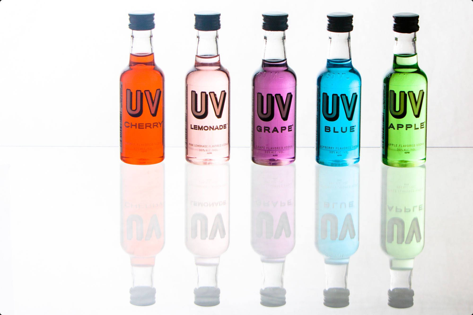 UV伏特加調味系列 壁紙 — 五款色彩繽紛的UV口味飲料瓶，包括櫻桃、檸檬水、葡萄、藍色和蘋果，排列在反光的白色表面上。