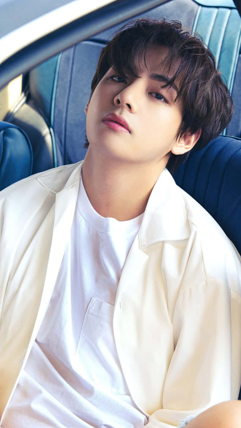 V BTS 休息臉手機壁紙 壁紙 — 一位深色頭髮的年輕男子穿著白色襯衫和外套，放鬆地坐在汽車的藍色皮革座椅上。