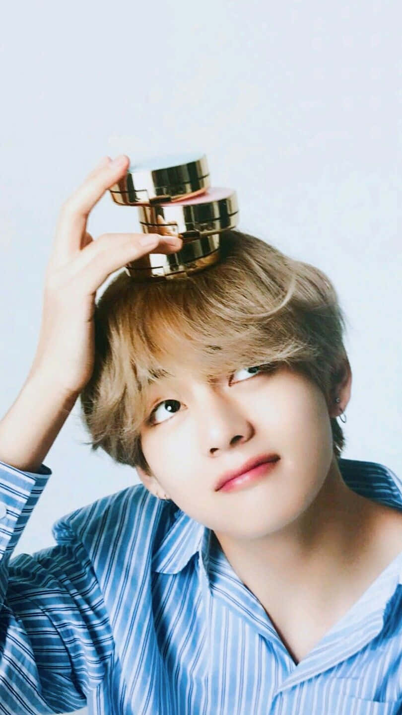 V BTS 手機微笑 壁紙 — 一位金髮年輕男子穿著藍色條紋襯衫，頭頂平衡著金色化妝品罐。