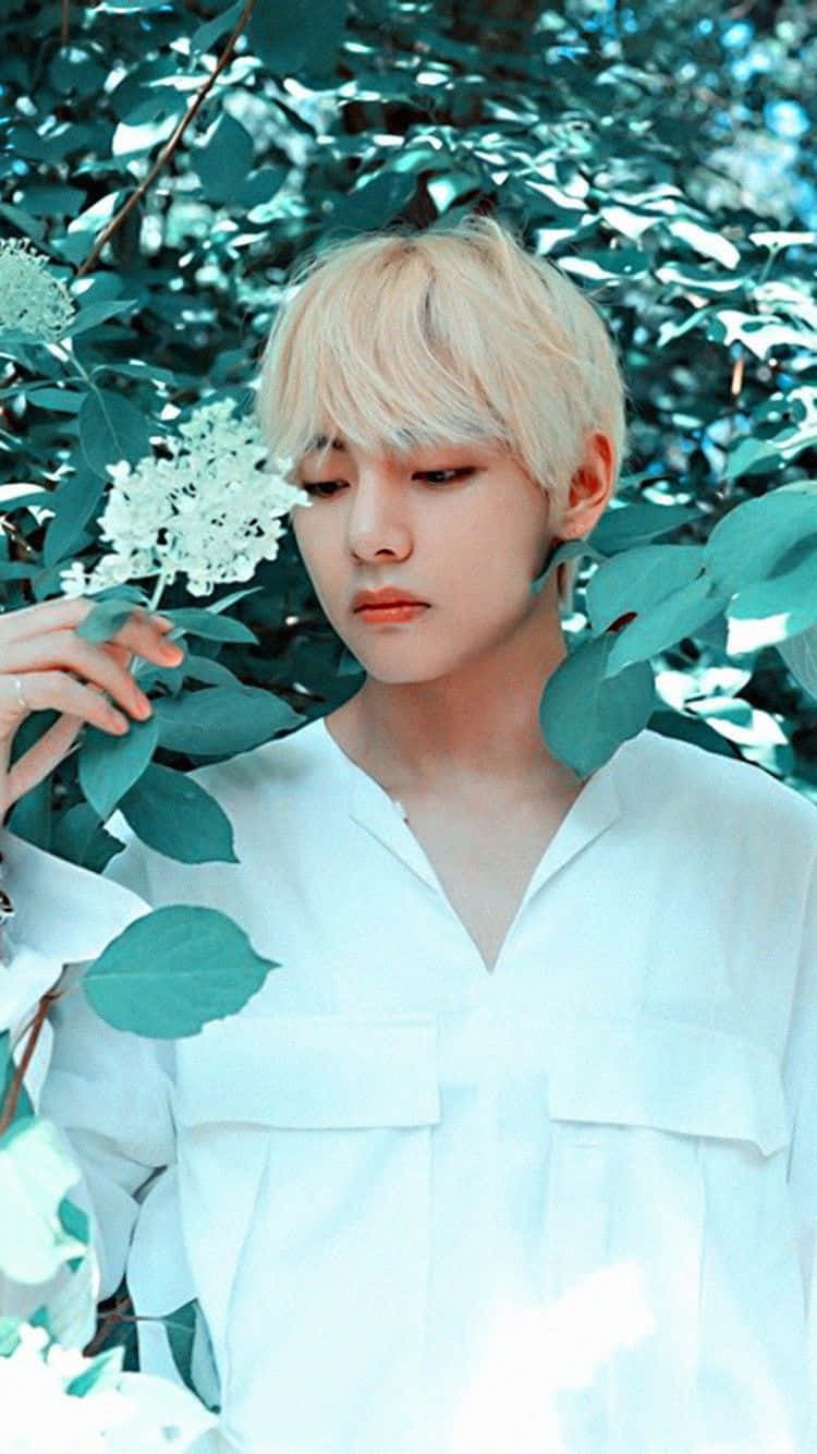 V BTS 手機花束風 壁紙 — 一位金髮年輕人穿著白襯衫，站在茂密的綠葉和白色花朵中，沐浴在柔和日光下。
