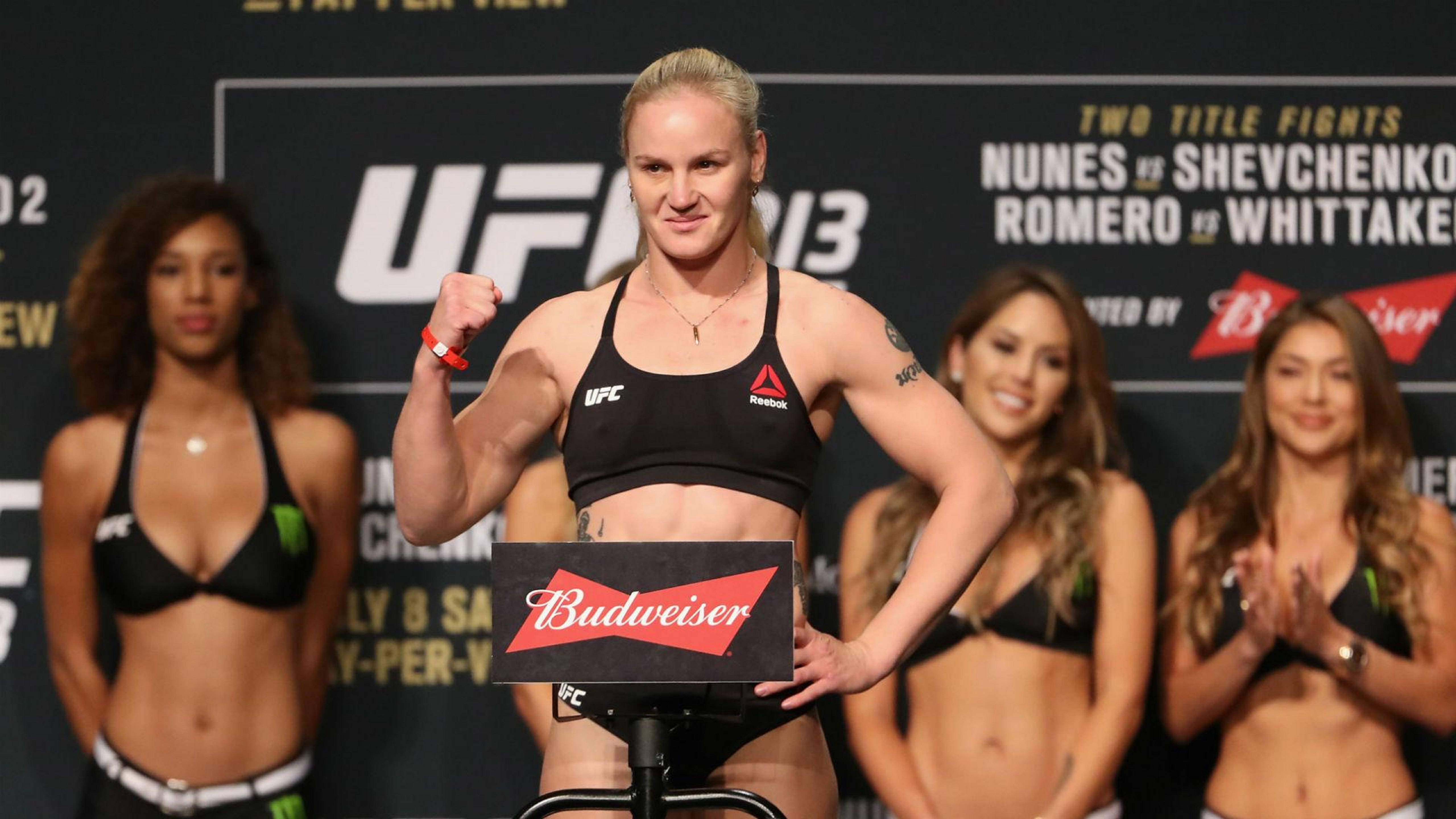 Valentina Shevchenko 称重 UFC 213 壁紙 — 一位女性UFC拳手在稱重活動中自信地擺姿勢，背景有宣傳橫幅和微笑的擂台女郎。