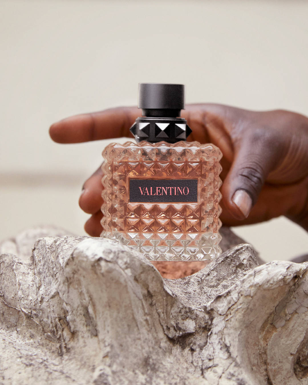 一隻手拿著 Valentino 香水瓶，瓶身帶有紋理玻璃，內有玫瑰金色液體。