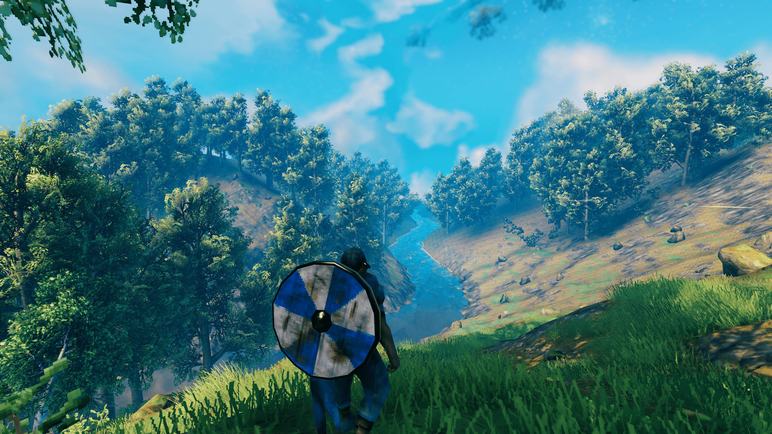 Valheim 武士的旅程 壁紙 — 一名戰士手持藍白盾牌，走過陽光明媚的草地山坡，俯瞰蜿蜒的河流，兩旁是茂密的森林。