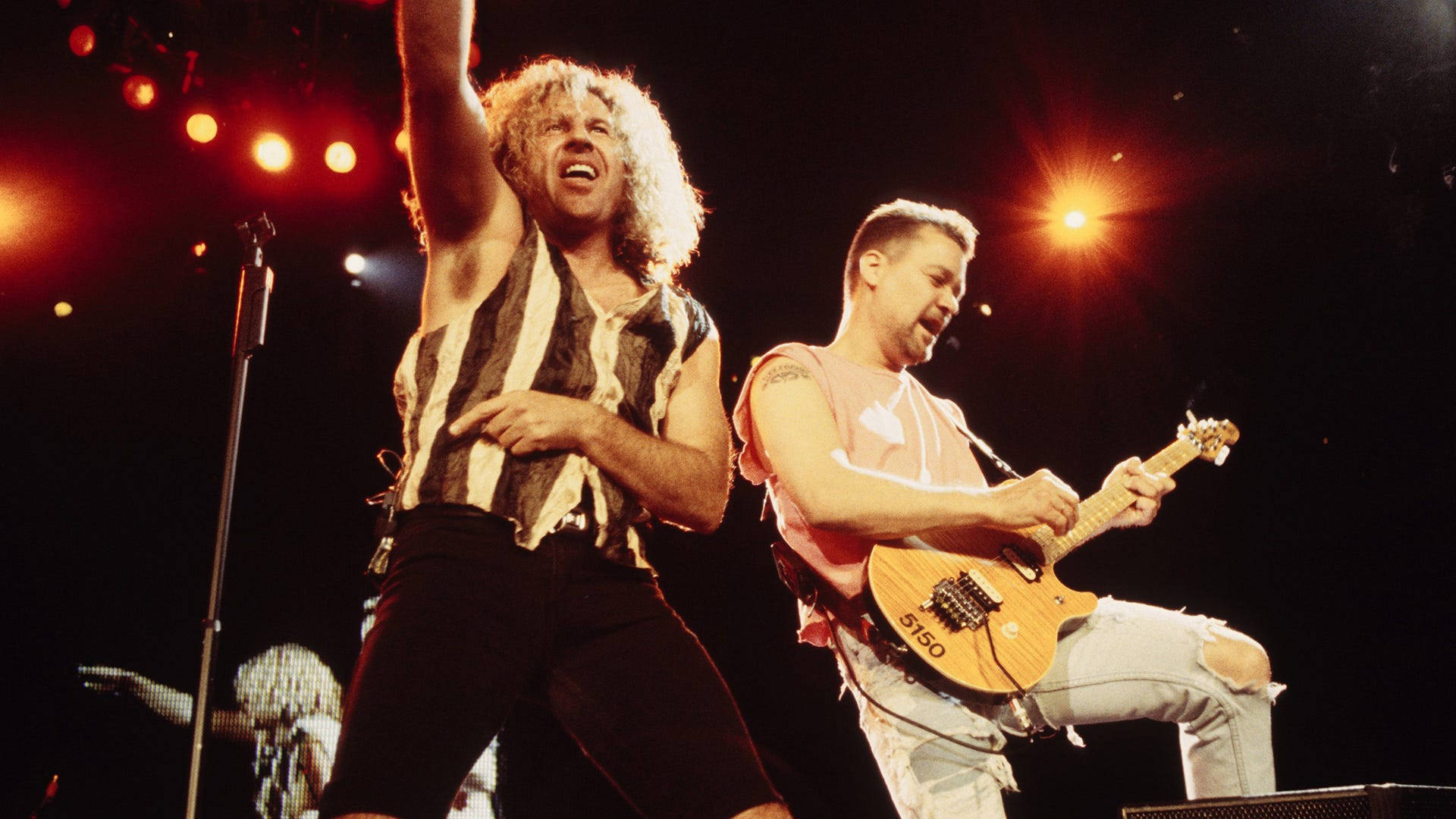 Van Halen 美國搖滾樂隊 壁紙 — 兩位搖滾音樂家在明亮的紅色聚光燈下充滿活力地表演，一位唱歌，另一位彈結他。