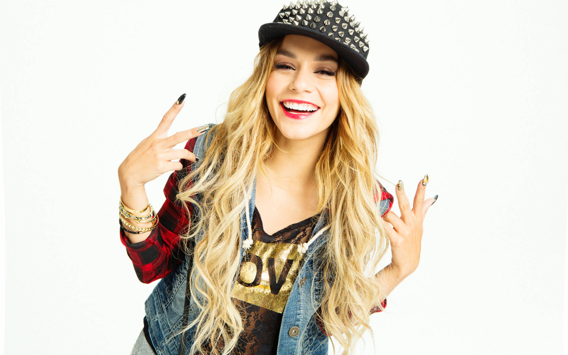 超級巨星 Vanessa Hudgens 壁紙 — 一位微笑的金髮女性戴著鉚釘帽和牛仔背心，在純白背景前做出搖滾手勢。