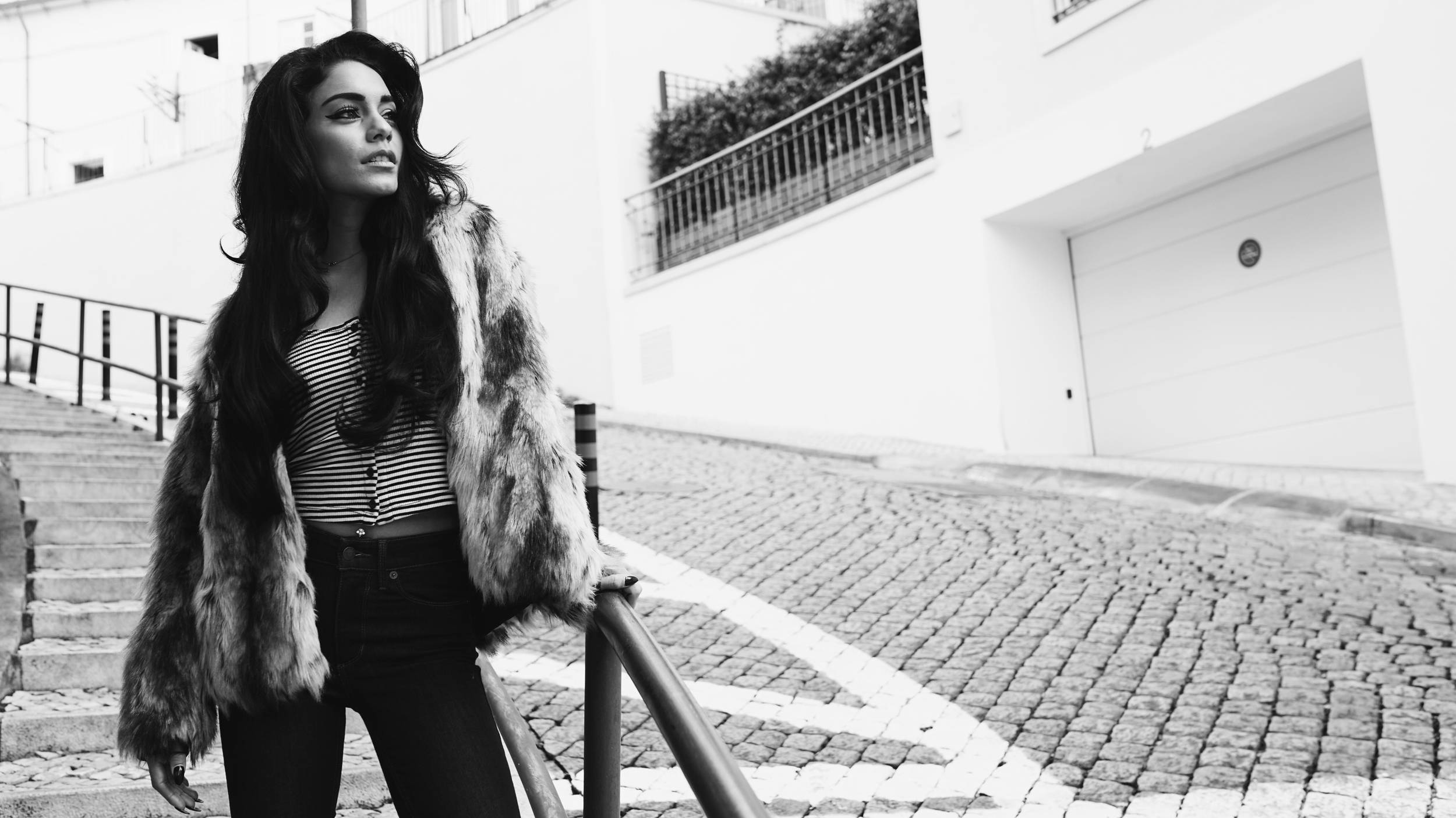 Vanessa Hudgens 黑白毛皮外套 壁紙 — 一位穿著毛皮大衣的女性自信地站在黑白相間的鵝卵石街道上，旁邊是樓梯和一棟白色建築。