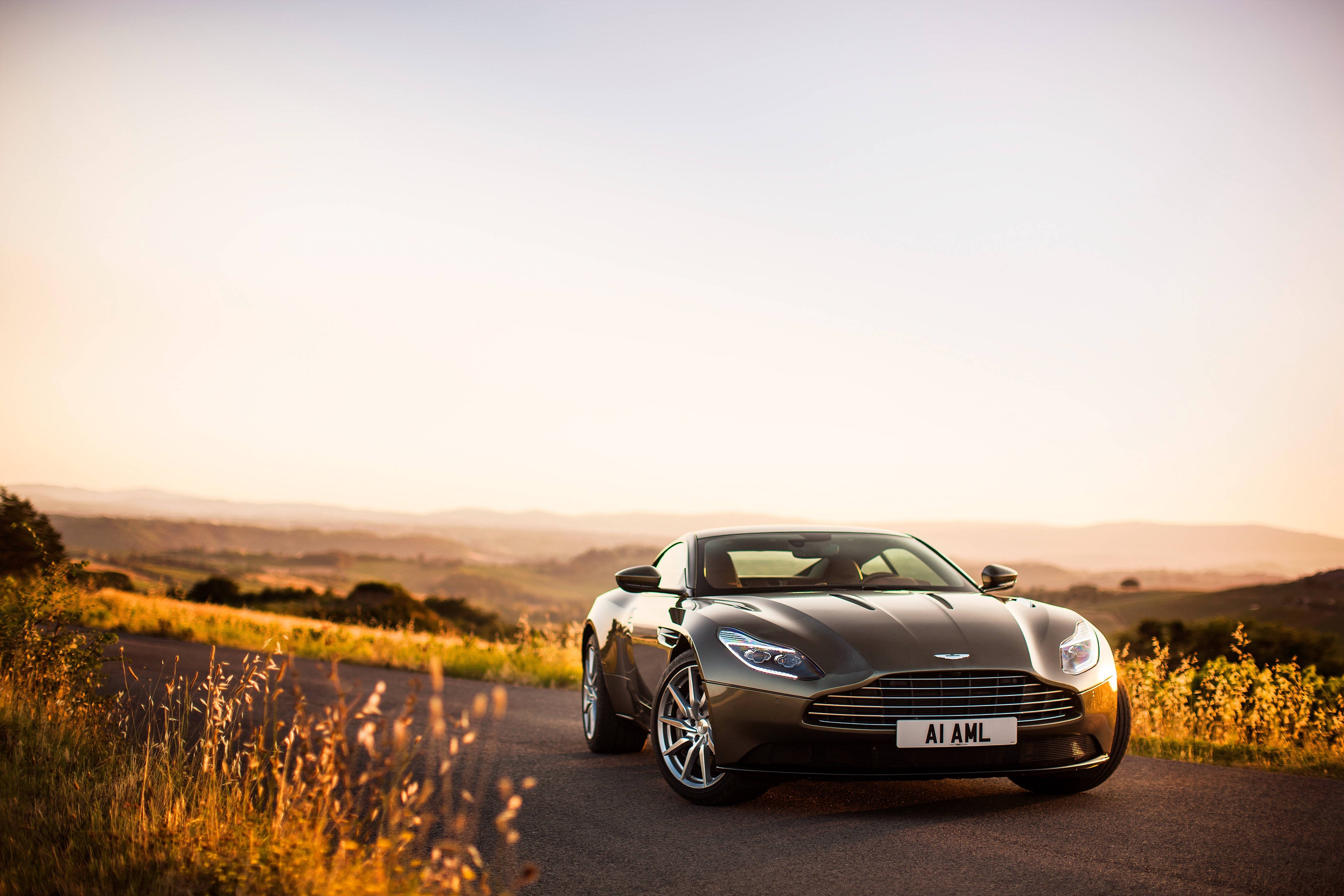 阿斯頓·馬丁 Vanquish 跑車 壁紙 — 一架流線型銀色Aston Martin DB11停泊在蜿蜒的鄉村道路上，背景是金色夕陽和起伏的山丘。