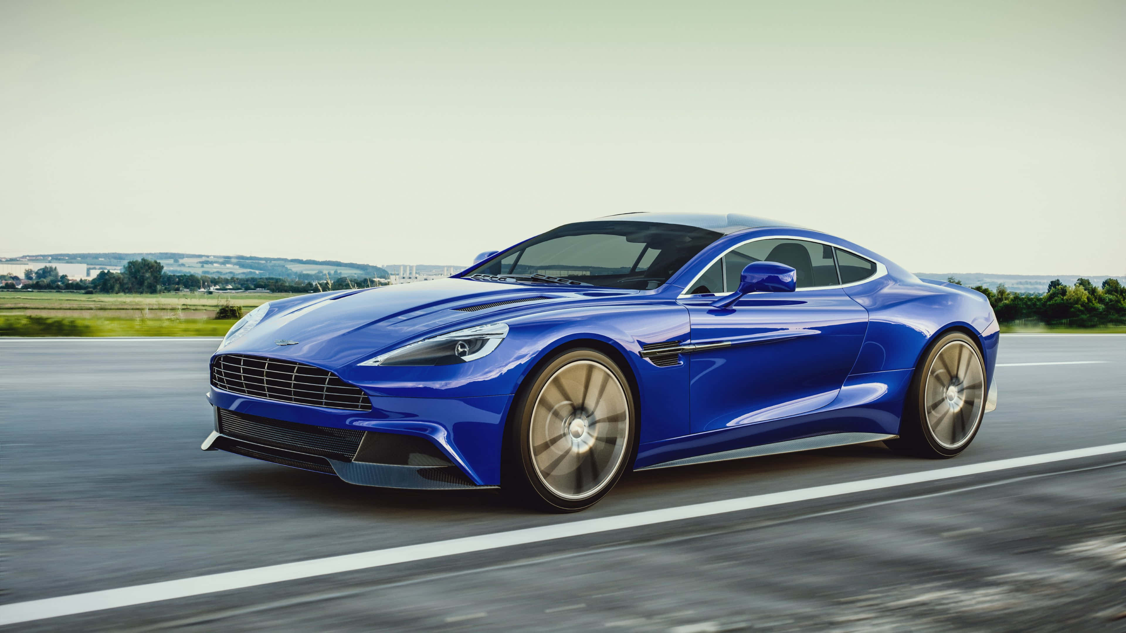 阿斯頓·馬丁 Vanquish S Ultimate – 象徵頂級豪華汽車體驗 壁紙 — 一架流線型的藍色Aston Martin跑車在柔和日光的天空下沿鄉村高速公路飛馳。