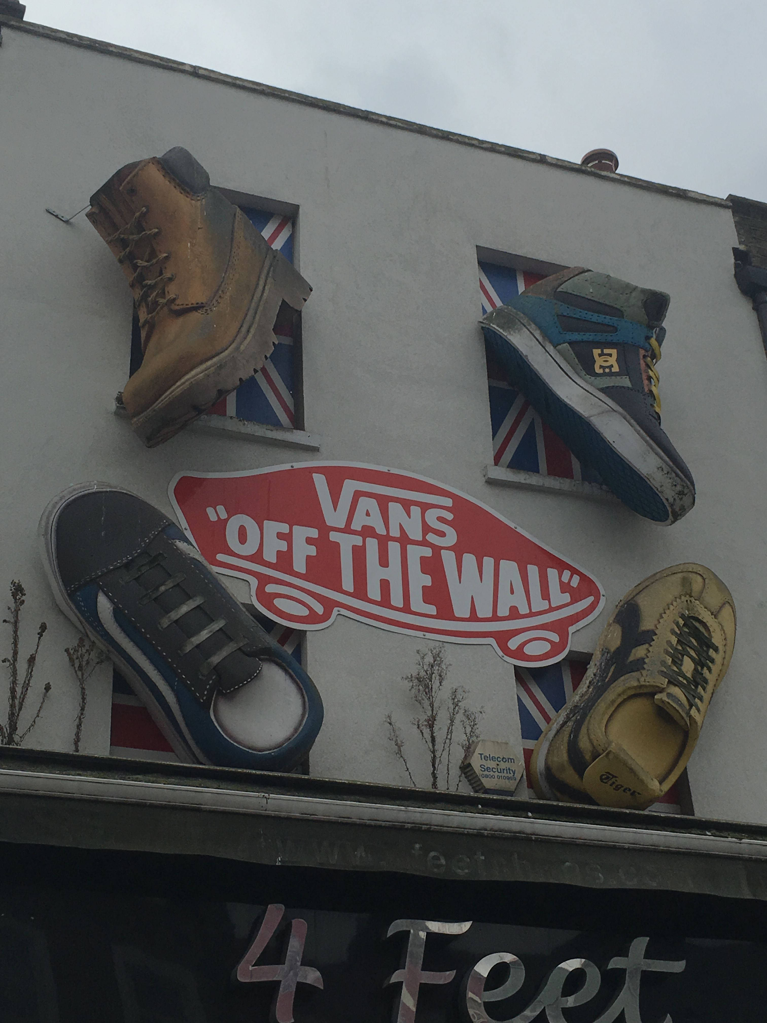 Vans Off The Wall 廣告牌 壁紙 — 四對復古運動鞋和一個Vans標誌安裝在建築物外牆上，窗框為英國國旗圖案，天空多雲。