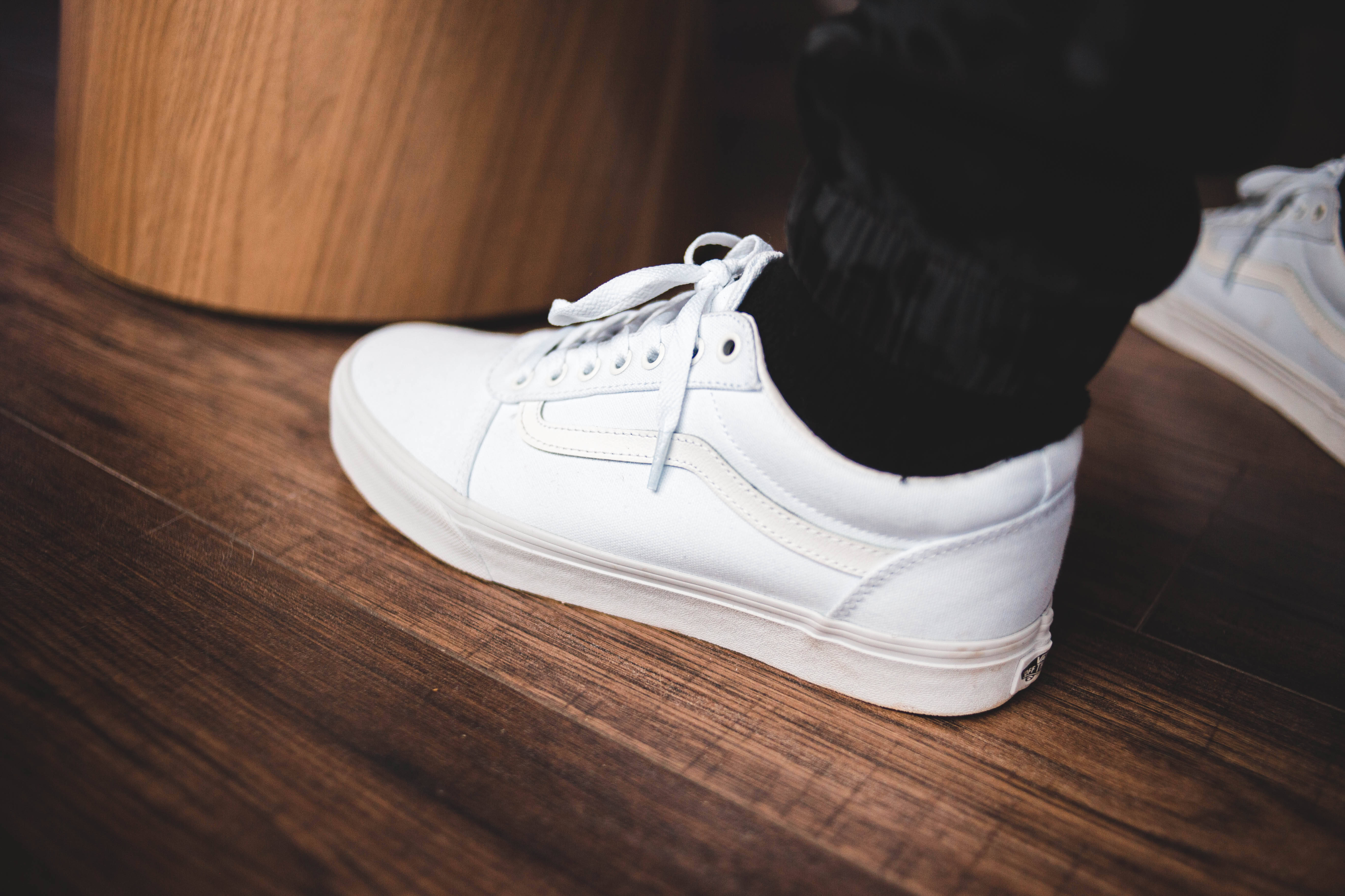 經典白色 Vans 運動鞋 壁紙 — 木地板上一隻白色運動鞋配黑色襪子，旁邊有一個木凳的近距離特寫。