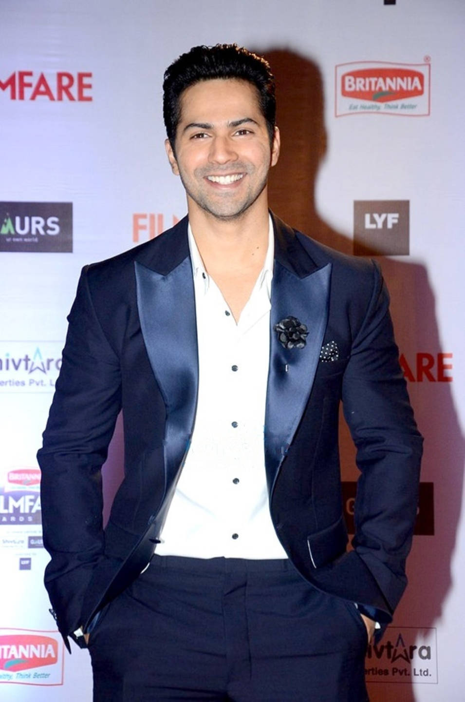 Varun Dhawan 出席2016年Filmfare獎典禮 壁紙 — 一位微笑的男子身穿海軍藍緞面翻領燕尾服外套和白色襯衫，在品牌活動背景前自信地擺姿勢。