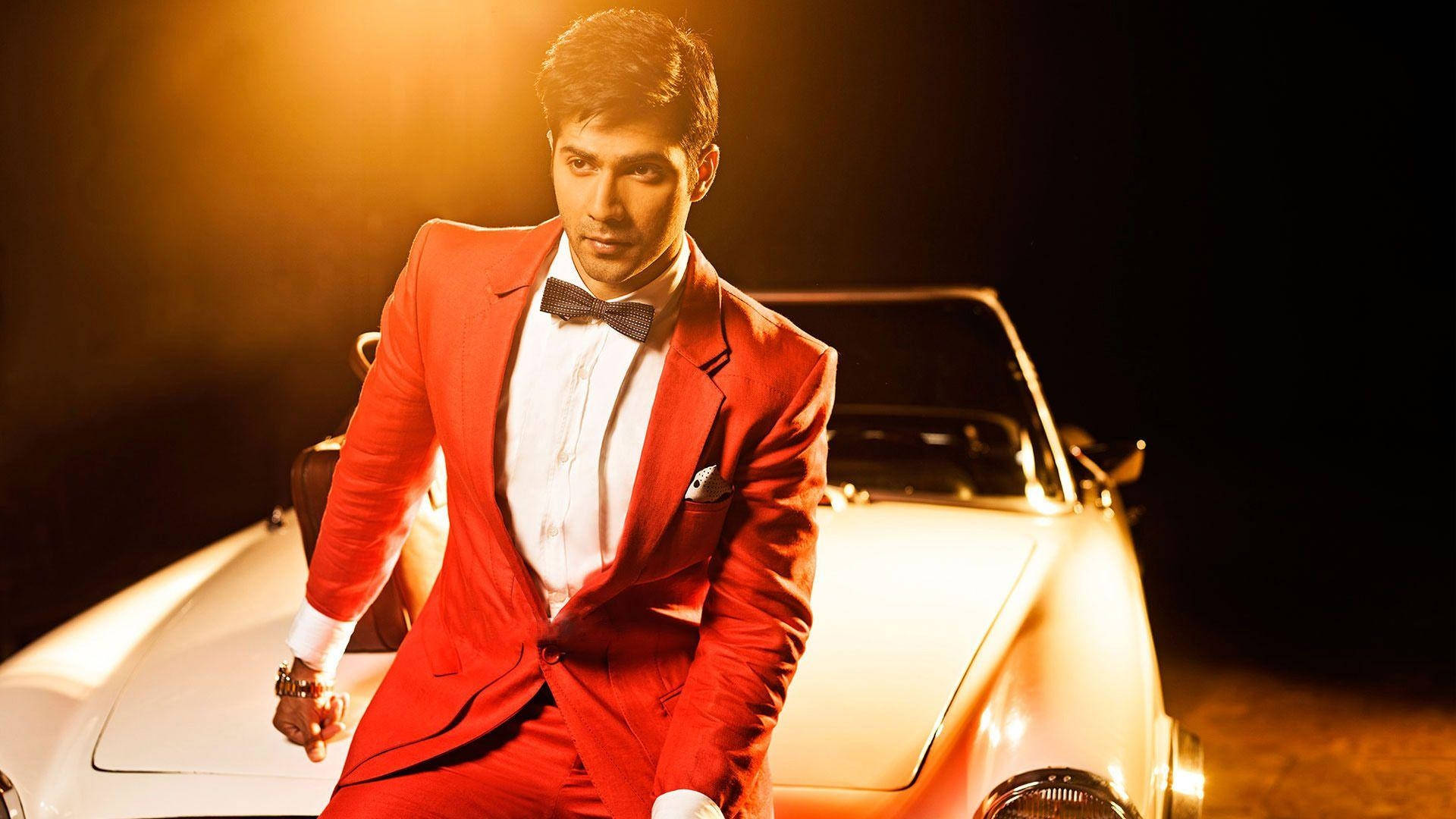 Varun Dhawan 坐於古董車上 壁紙 — 一名身穿搶眼紅色西裝的男子，在戲劇性的金色燈光下自信地靠在一輛白色跑車上。