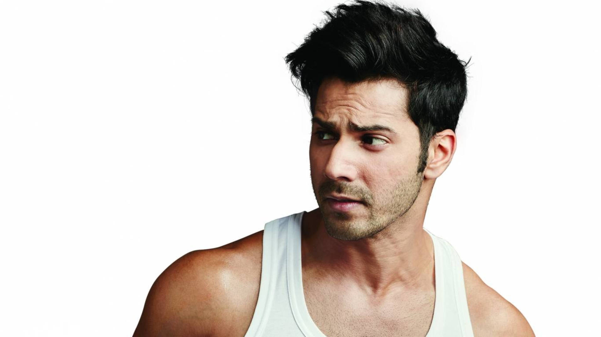 Varun Dhawan 穿白色背心 壁紙 — 一個黑髮帶鬚根的男人穿著白色背心，在純白背景前側目而視，表情帶點懷疑。