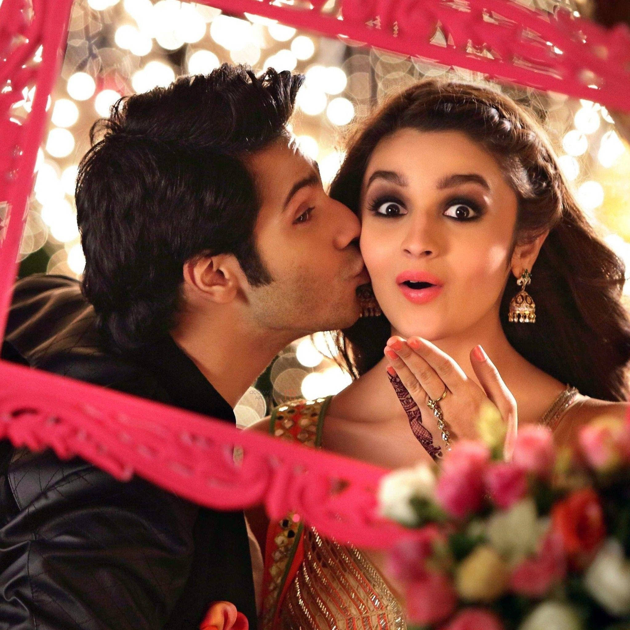Varun Dhawan 與 Alia Bhatt 壁紙 — 一個男人親吻一個驚訝的女人的臉頰，周圍是粉紅色裝飾邊框，背後有發光的燈光。