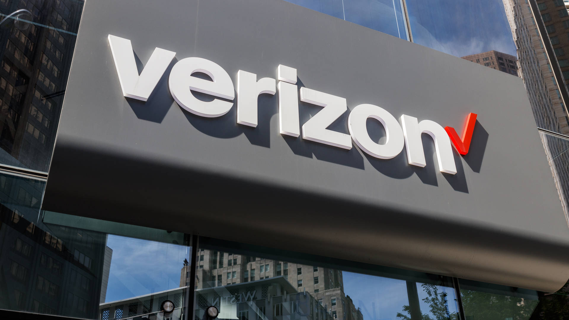 Verizon — 移動網絡技術領先者 壁紙 — 一個大型灰色Verizon標誌，帶有白色字體和紅色剔號，安裝在玻璃幕牆建築上，反射出城市摩天大樓。