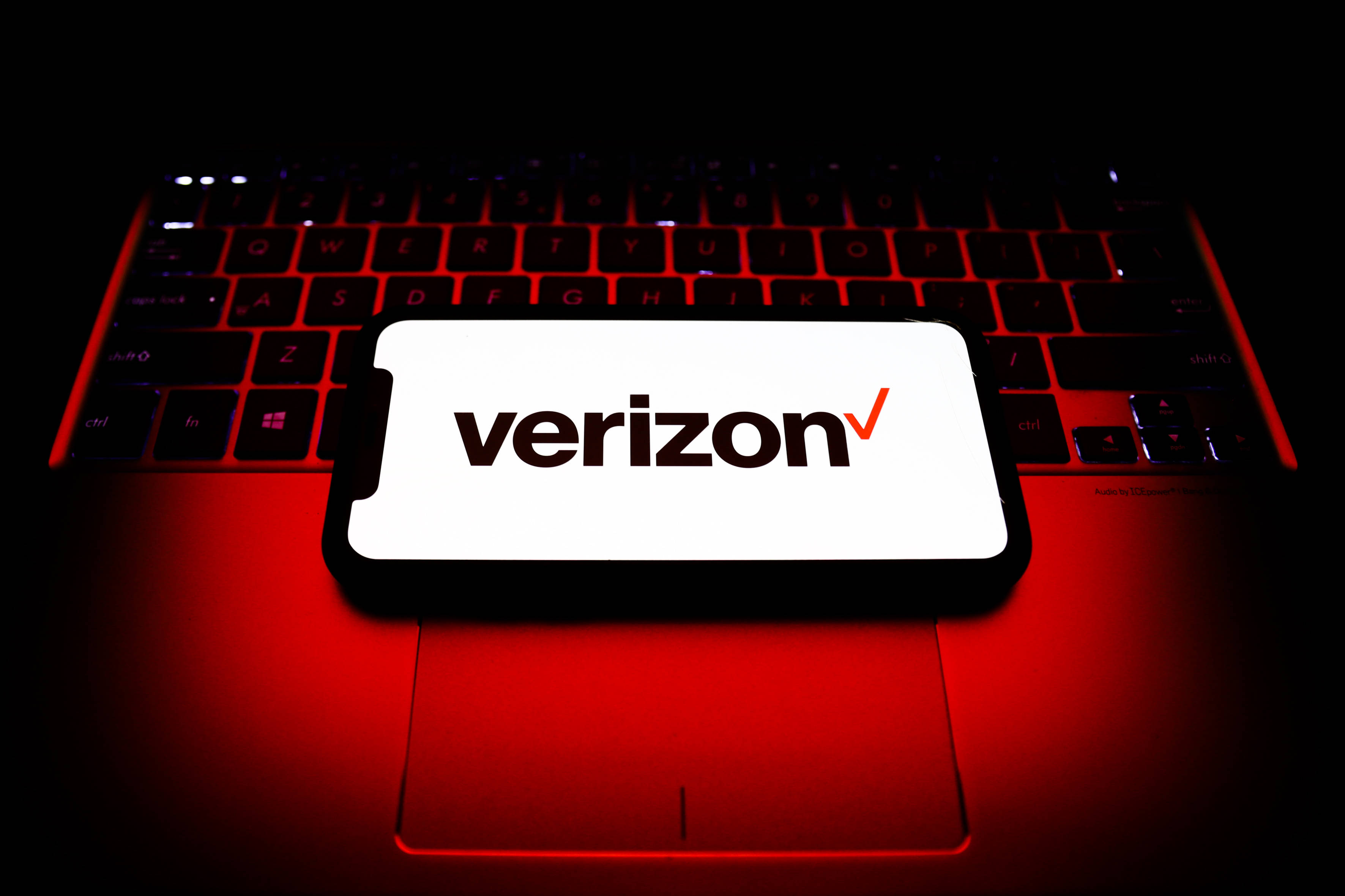 Verizon iPhone 優惠方案 壁紙 — 一部顯示Verizon標誌的智能手機放在一個被強烈紅色背光照亮的筆記型電腦鍵盤上，背景為深色。