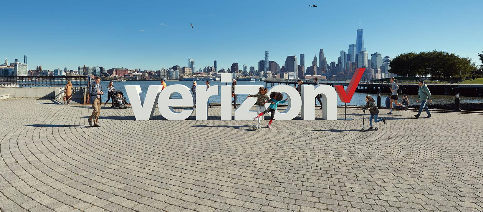 Verizon SLU 標誌 壁紙 — 人們在明亮的藍天和紐約市天際線背景下，圍繞著一個帶有紅色勾號的巨大白色Verizon標誌漫步和玩耍。