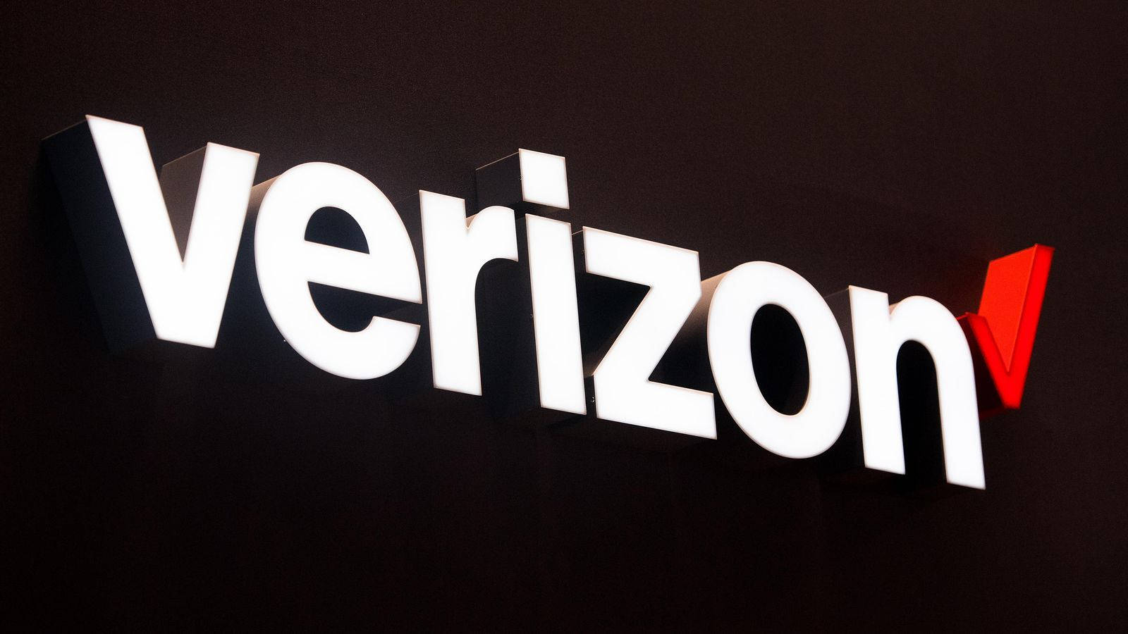 Verizon 電信公司標誌 壁紙 — 一個明亮照明的白色和紅色Verizon標誌招牌在現代室內環境中的深色牆壁上脫穎而出。