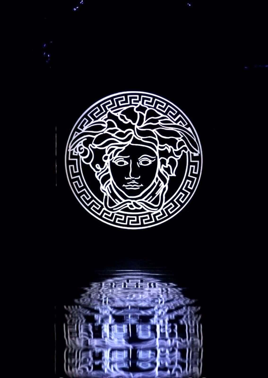 Versace iPhone 水面倒影標誌 壁紙 — 一個發光的白色美杜莎頭標誌，帶有希臘鑰匙邊框，漂浮在黑暗水面上扭曲的藍色倒影之上。