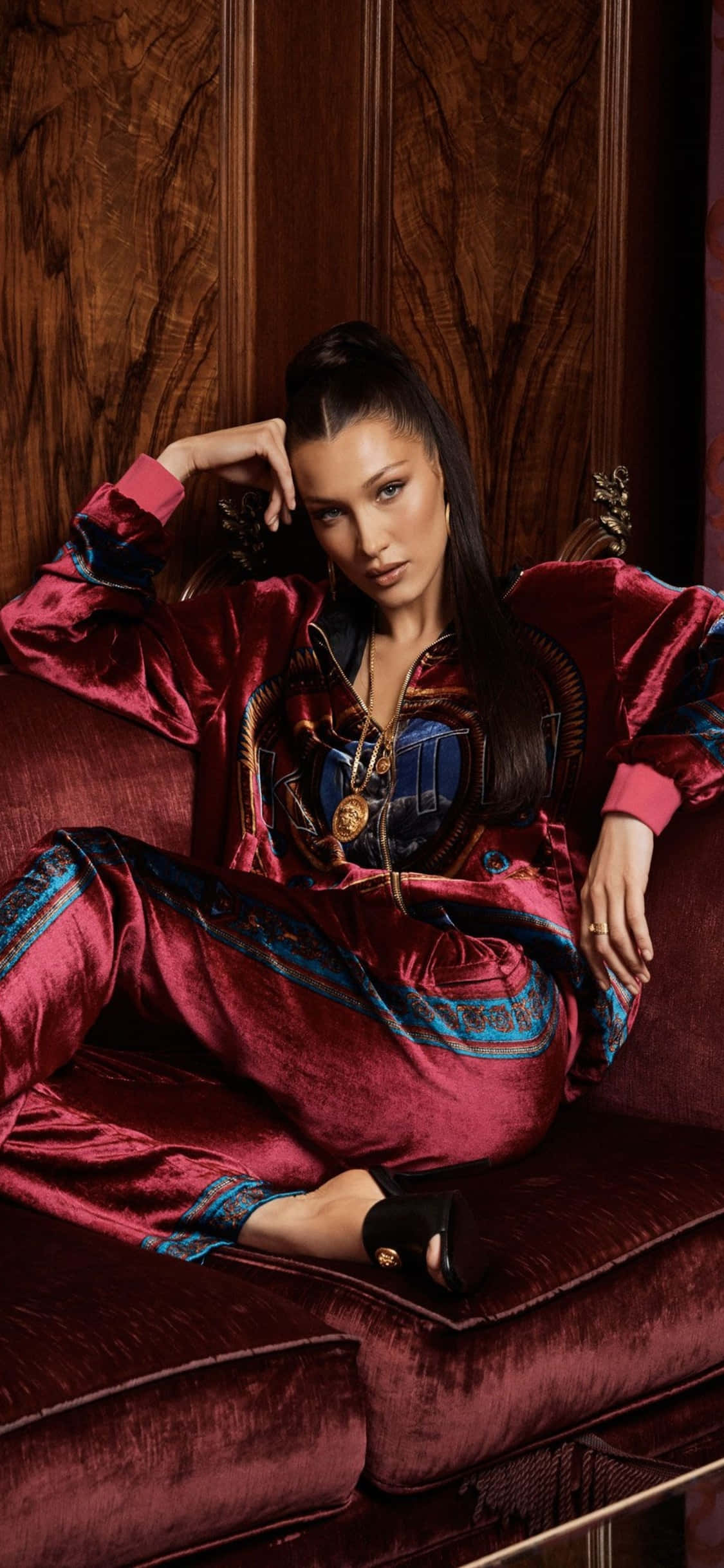 Versace iPhone 貝拉·哈迪德紅絨連身裝 壁紙 — 一位身穿華麗紅色天鵝絨套裝的女性，自信地躺在一張相配的天鵝絨沙發上，背景是深色木質牆板。