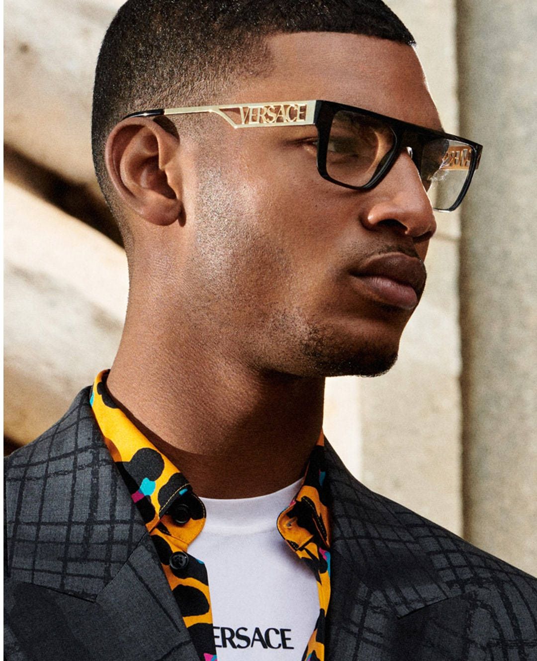 Versace VE3326U 眼鏡男性模特 壁紙 — 一位時尚男士戴著大膽的 Versace 太陽眼鏡，穿著充滿活力的圖案襯衫，外搭格紋西裝外套，背景是紋理石牆。