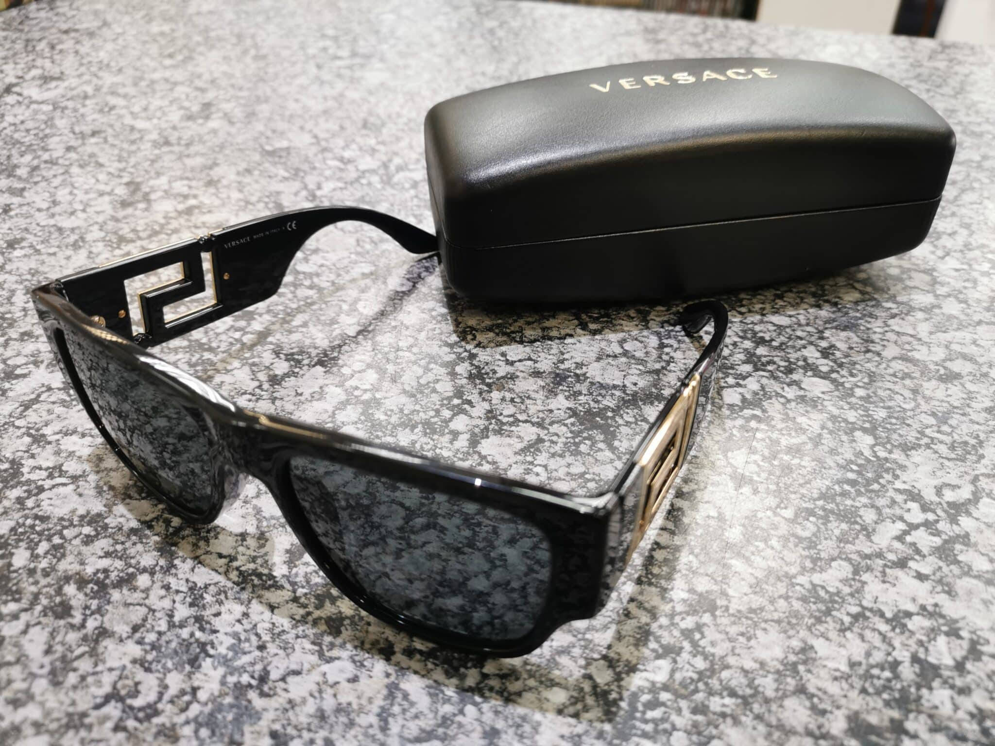 Versace VE4296 女士太陽眼鏡 壁紙 — 黑色Versace太陽眼鏡帶金色標誌細節，旁邊是其品牌眼鏡盒，放在斑點灰色檯面上。