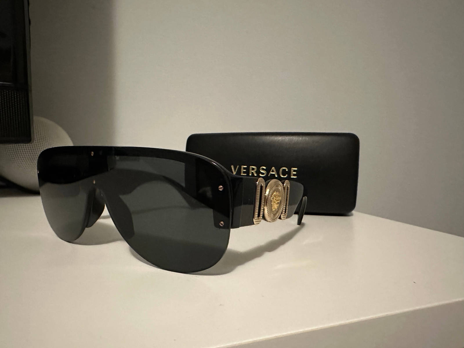 Versace 中性款盾形/飛行員太陽眼鏡 4391 壁紙 — 帶有金色美杜莎標誌的黑色Versace太陽眼鏡大膽地放在白色表面上，旁邊是其品牌眼鏡盒。