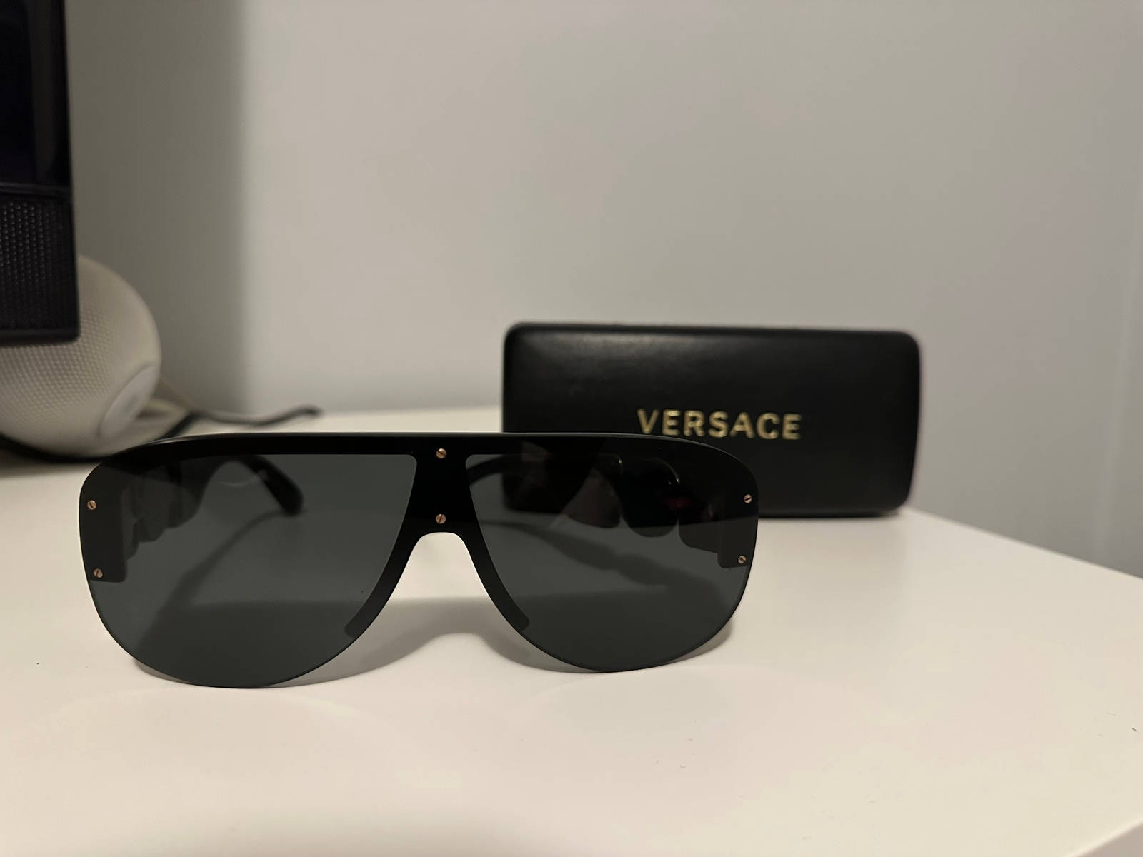 Versace 太陽眼鏡 VE 4391 壁紙 — 帶有金色鉚釘的醒目黑色Versace太陽眼鏡，放在白色表面上，旁邊是其品牌眼鏡盒。