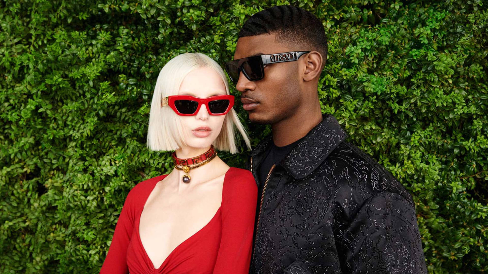 Versace 秋冬兩位模特 壁紙 — 一對時尚情侶在茂密的綠色籬笆前擺姿勢，戴著大膽的紅色和黑色太陽眼鏡，穿著時尚服裝。