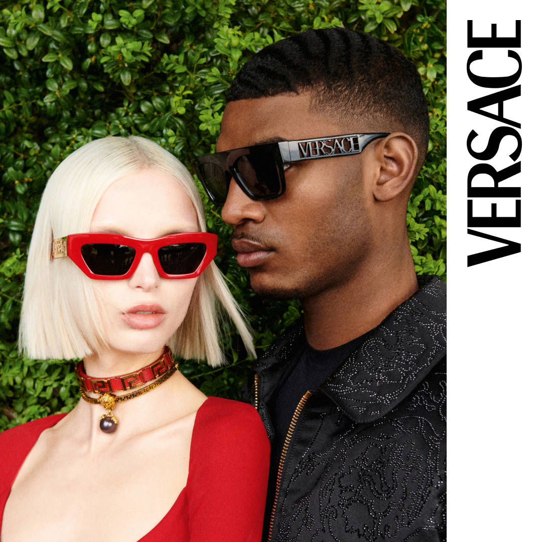 Versace秋冬系列模特展示 壁紙 — 一對時尚情侶戴著大膽的紅色和黑色 Versace 太陽眼鏡，在茂密的綠色樹籬背景下擺姿勢。