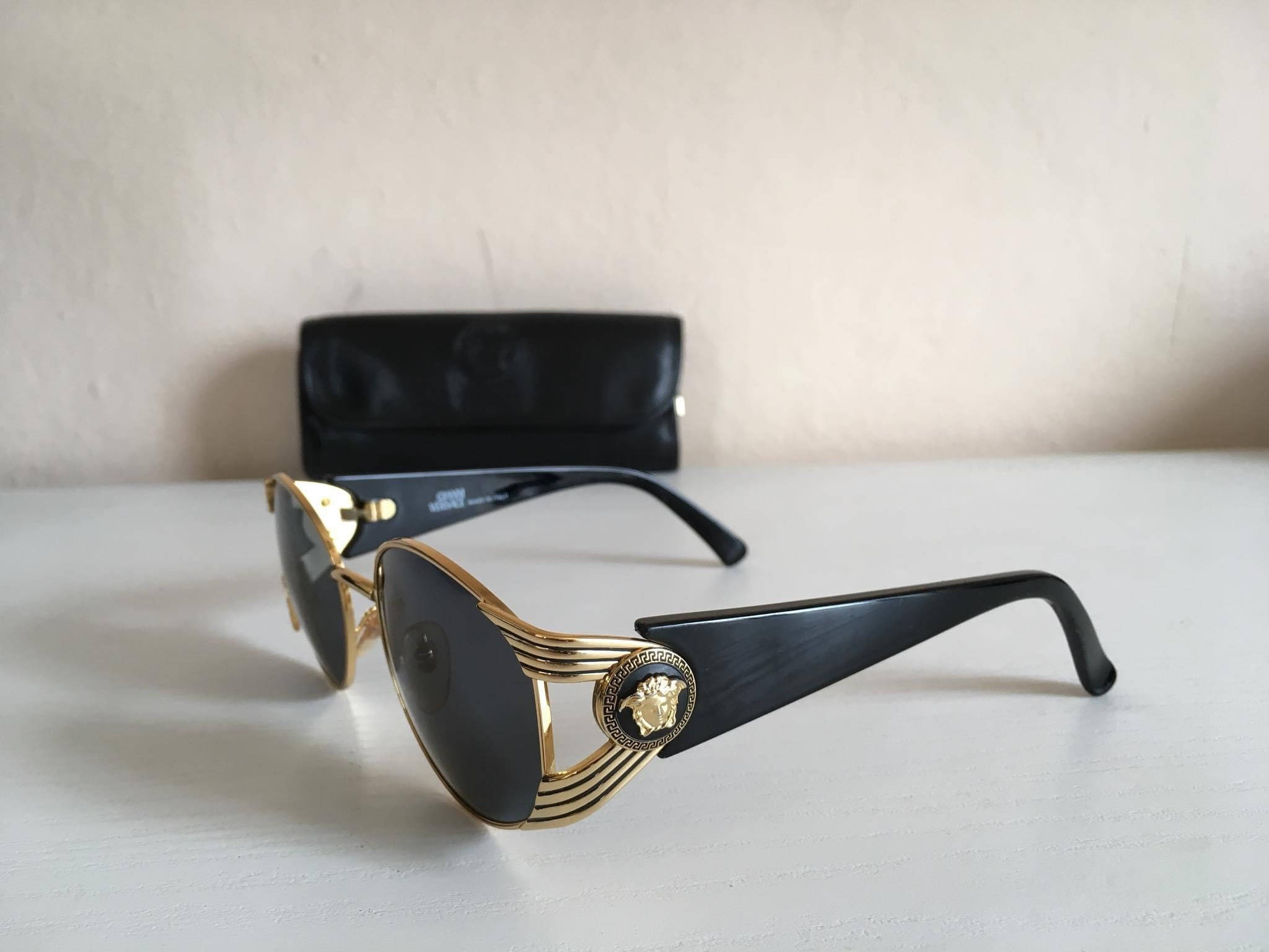 Versace金色復古太陽眼鏡 壁紙 — 優雅的金色和黑色 Versace 太陽眼鏡，帶有美杜莎標誌，放置在白色表面上，旁邊有一個黑色眼鏡盒。