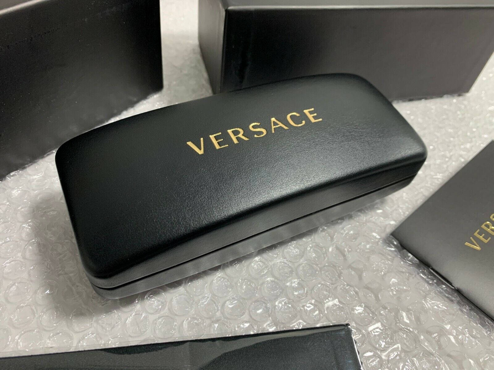 Versace黑色蚌殼鏡盒 壁紙 — 一個時尚的黑色Versace太陽眼鏡盒，帶有金色字樣，放在氣泡包裝上，旁邊是相配的盒子。