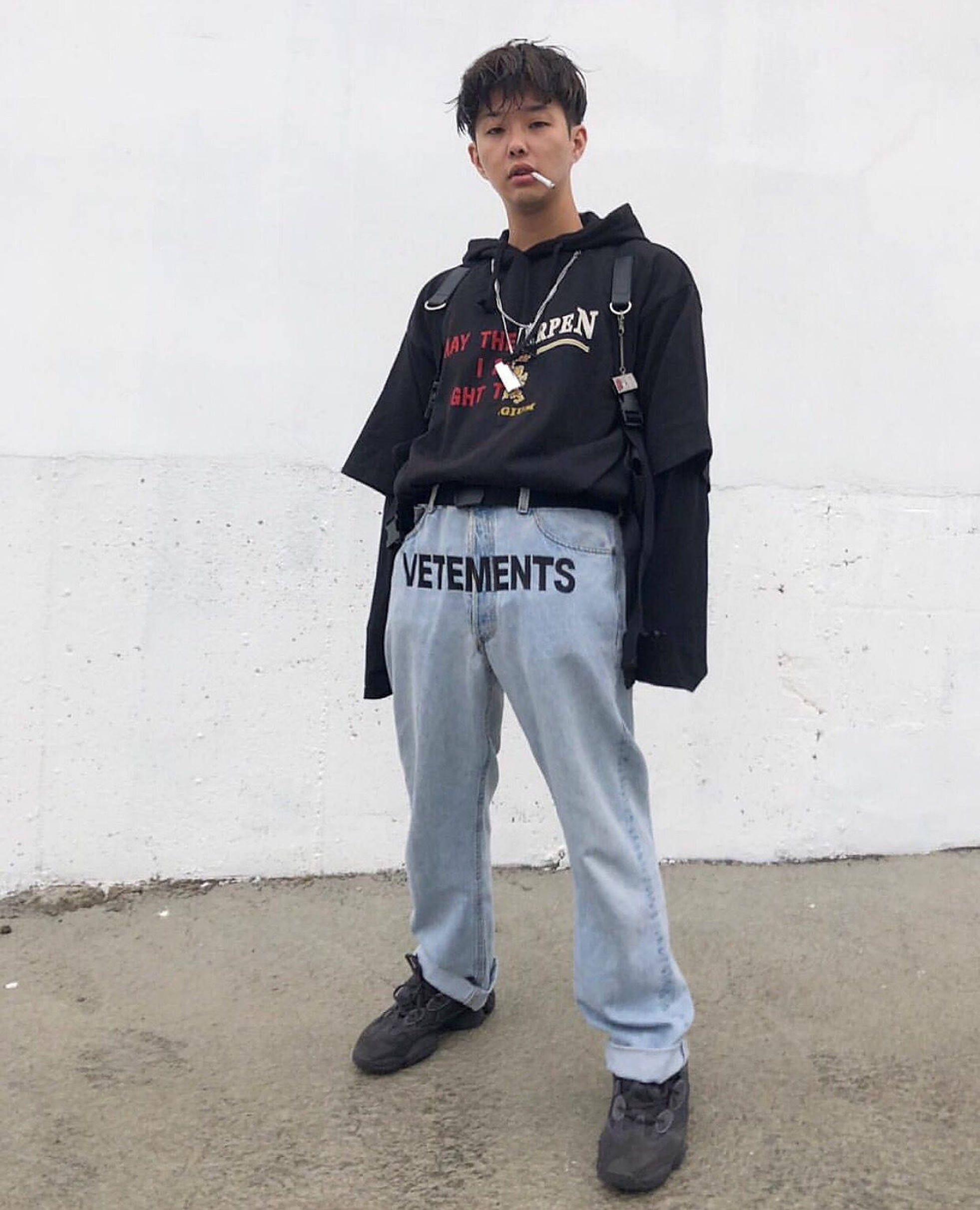 Vetements 牛仔褲 壁紙 — 一個穿著黑色連帽衫和名牌牛仔褲的年輕男子靠著純白牆壁，手持香煙。